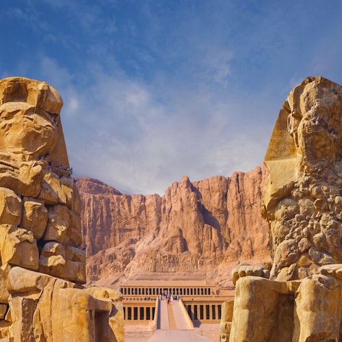 Ancient Egyptian temple entrance flanked by two large stone statues with mountainous desert landscape in the background.