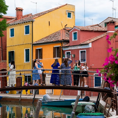 游客站在运河上的一座小桥上拍照，背景中是色彩缤纷的建筑和一艘小船。