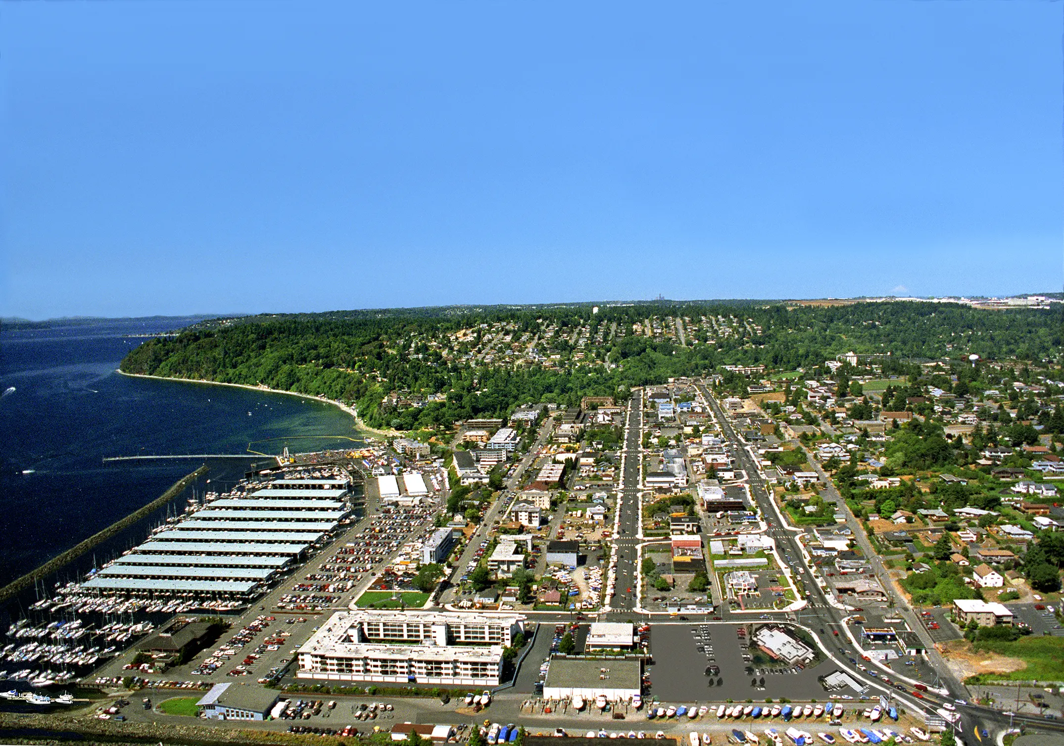 An aerial image of the City of Des Moines. The Puget Sound is to the left, roads and a cross-street grid in the center, and more forested areas to the right and farther in the background to the city.