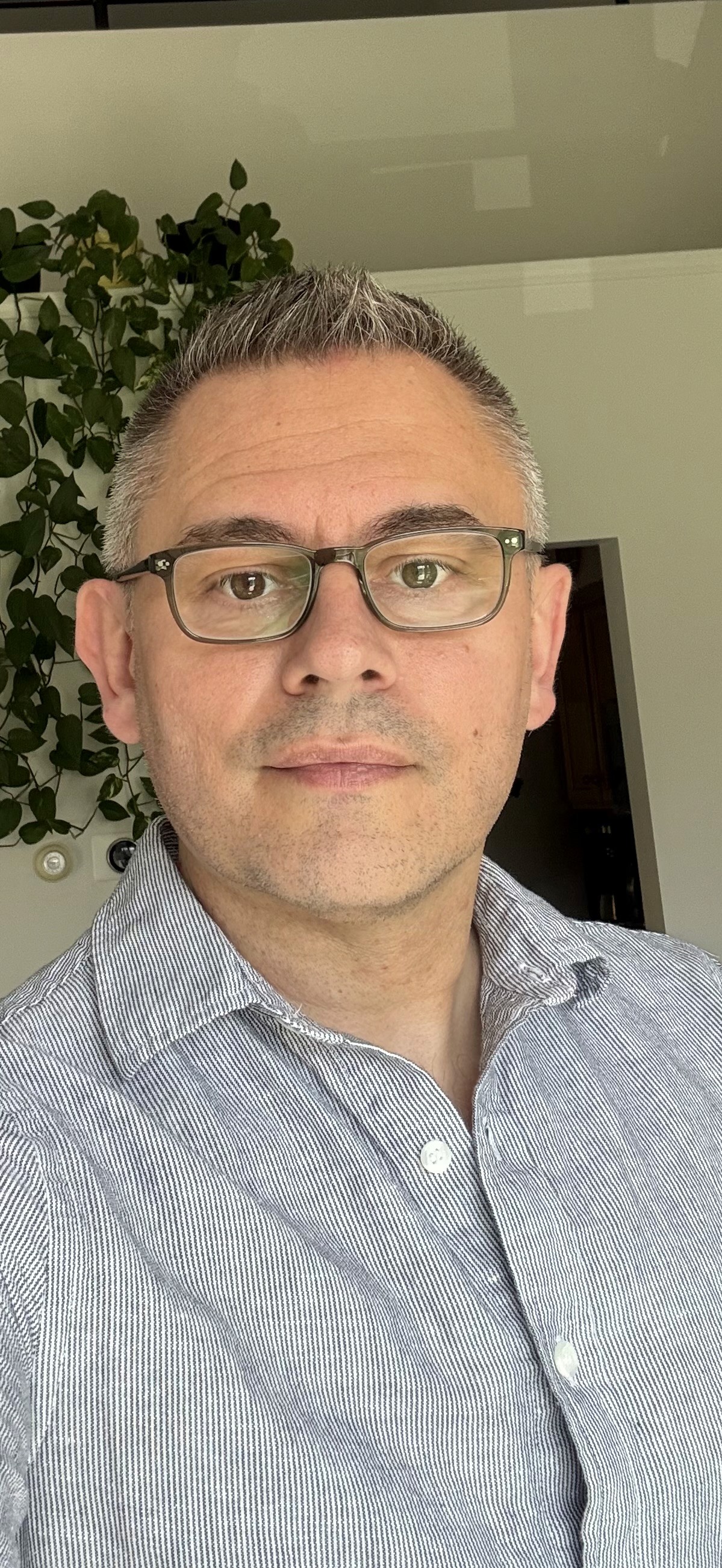 A person wearing glasses and a light gray button-up shirt stands indoors, with green plant leaves visible in the background.