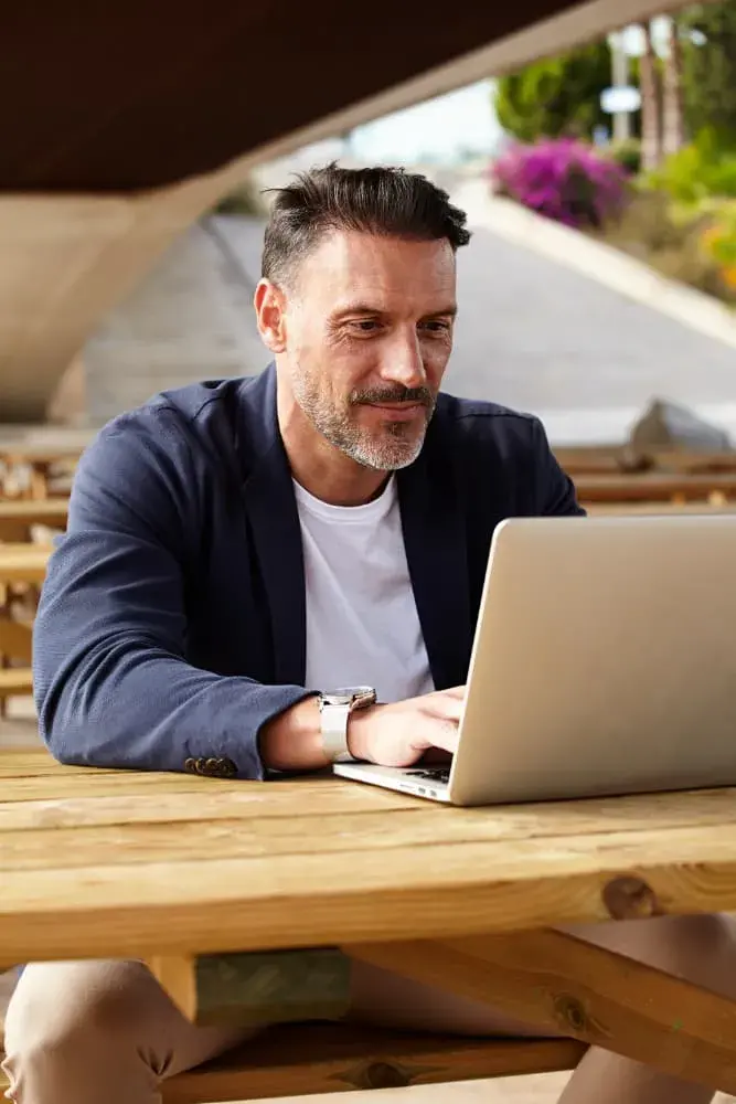 A man works on his laptop at an outdoor wooden table, dressed in a blazer and casual shirt. The image represents learning and researching real estate investment options, aligning with the educational resources provided by Chris Lewis Home Loans.