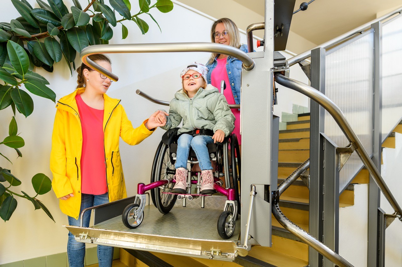 A young girl in a wheelchair is on a stairlift, smiling and holding hands with a woman in a yellow coat. Another woman stands behind her, ensuring her safety. They are in a bright indoor area near stairs and a large plant.