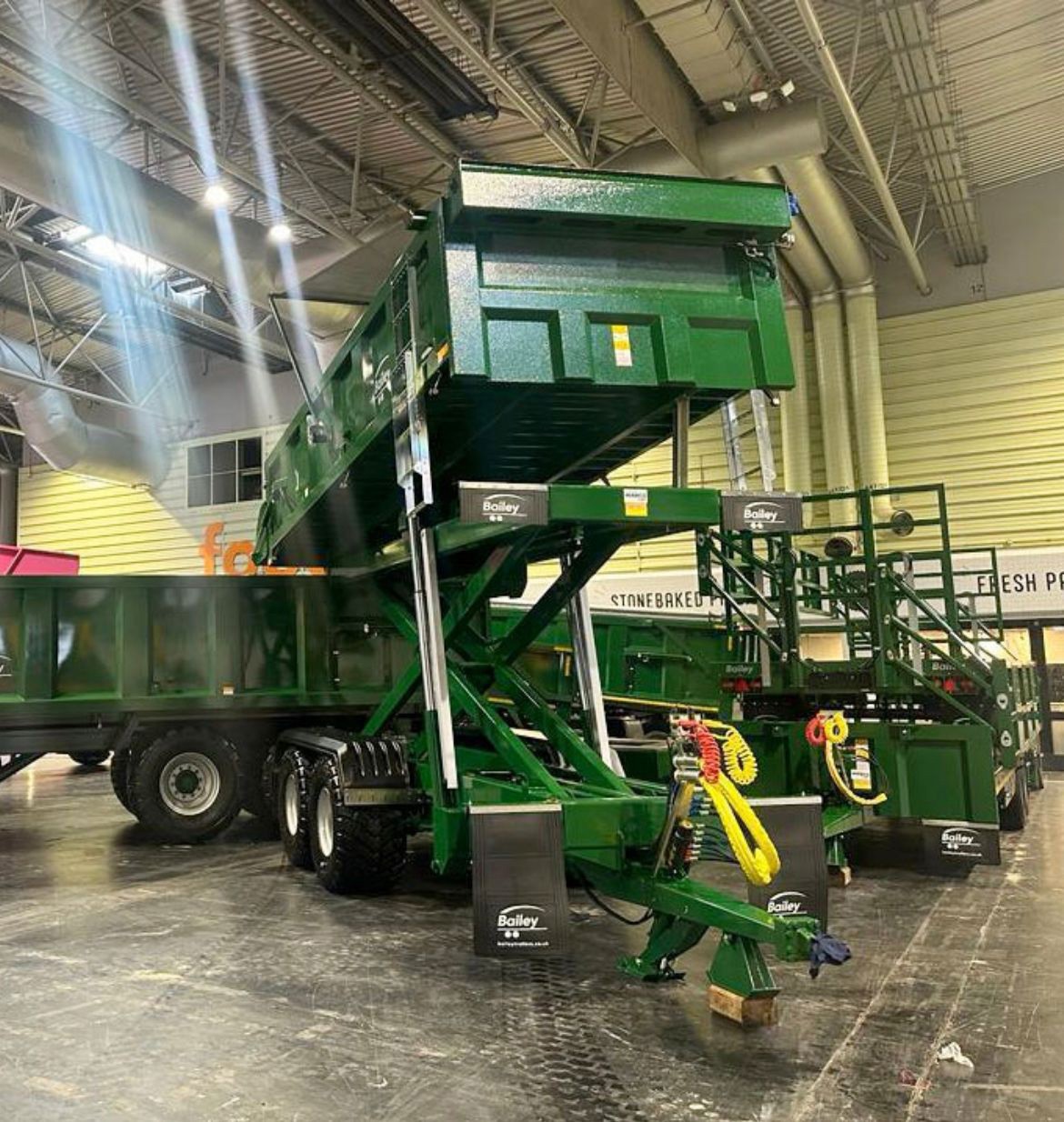 A green Bailey hydraulic tipping trailer with raised scissor-lift mechanism on display inside an agricultural trade show exhibition hall.