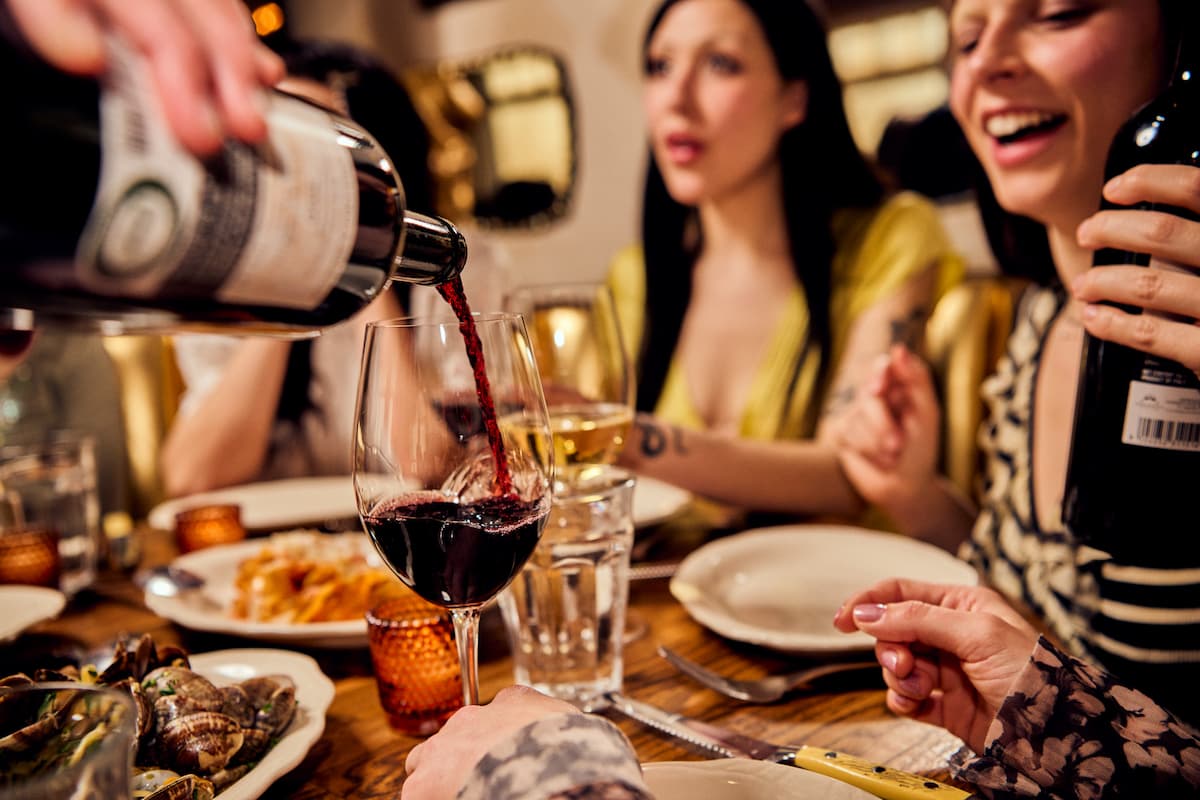 A server pouring red wine for a group of friends seated at a restaurant table.