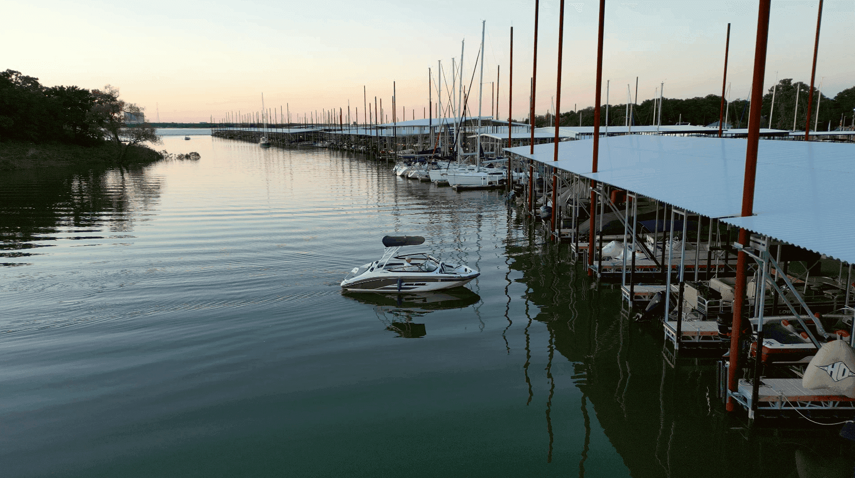 A serene marina scene at sunset, featuring numerous sailboats docked at covered slips, with calm water reflecting the warm colors of the sky.