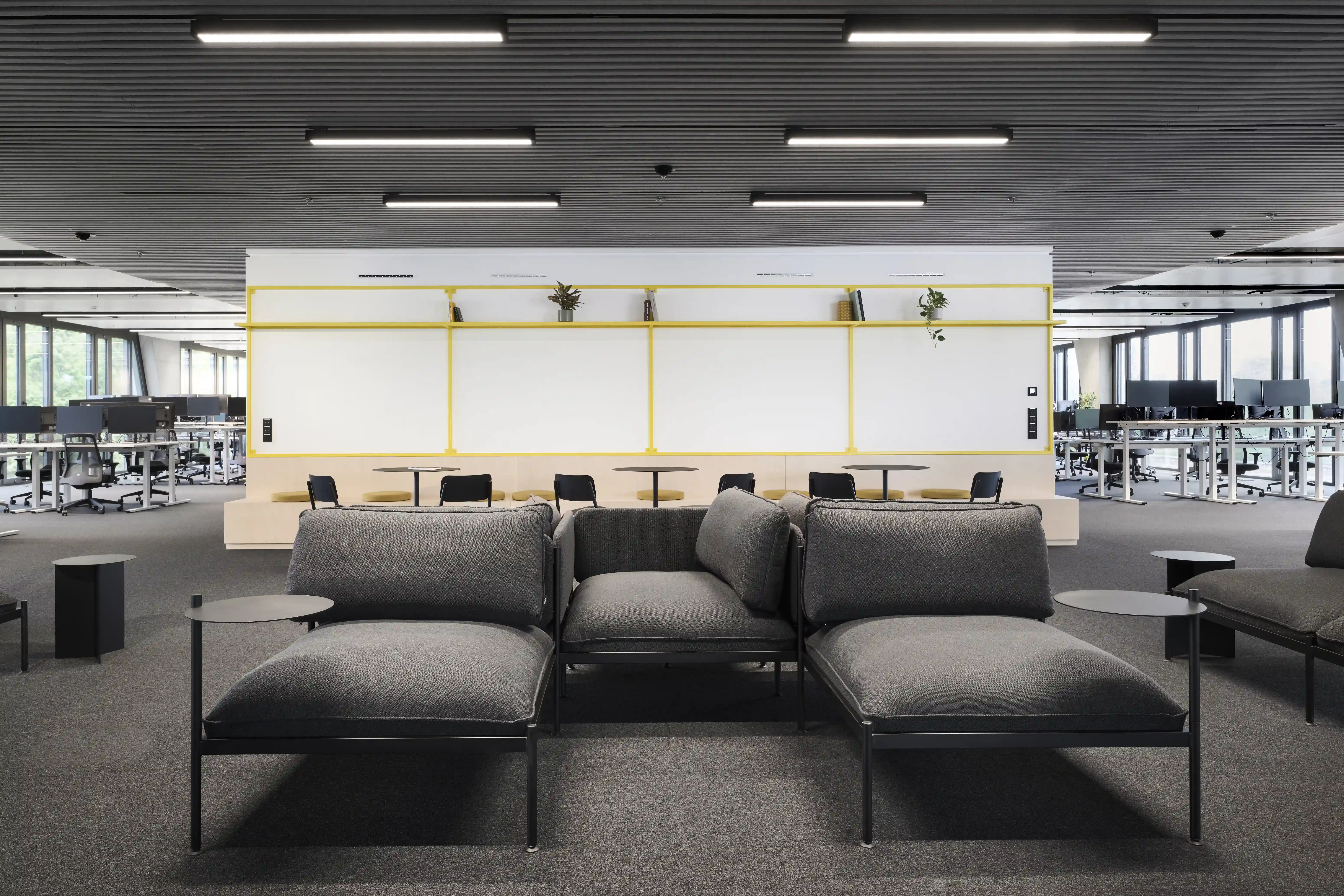 Open-plan office at ENPAL with a grey modular sofa group and black tray tables in the centre, whiteboard walls with yellow frames and rows of height-adjustable desks beyond.