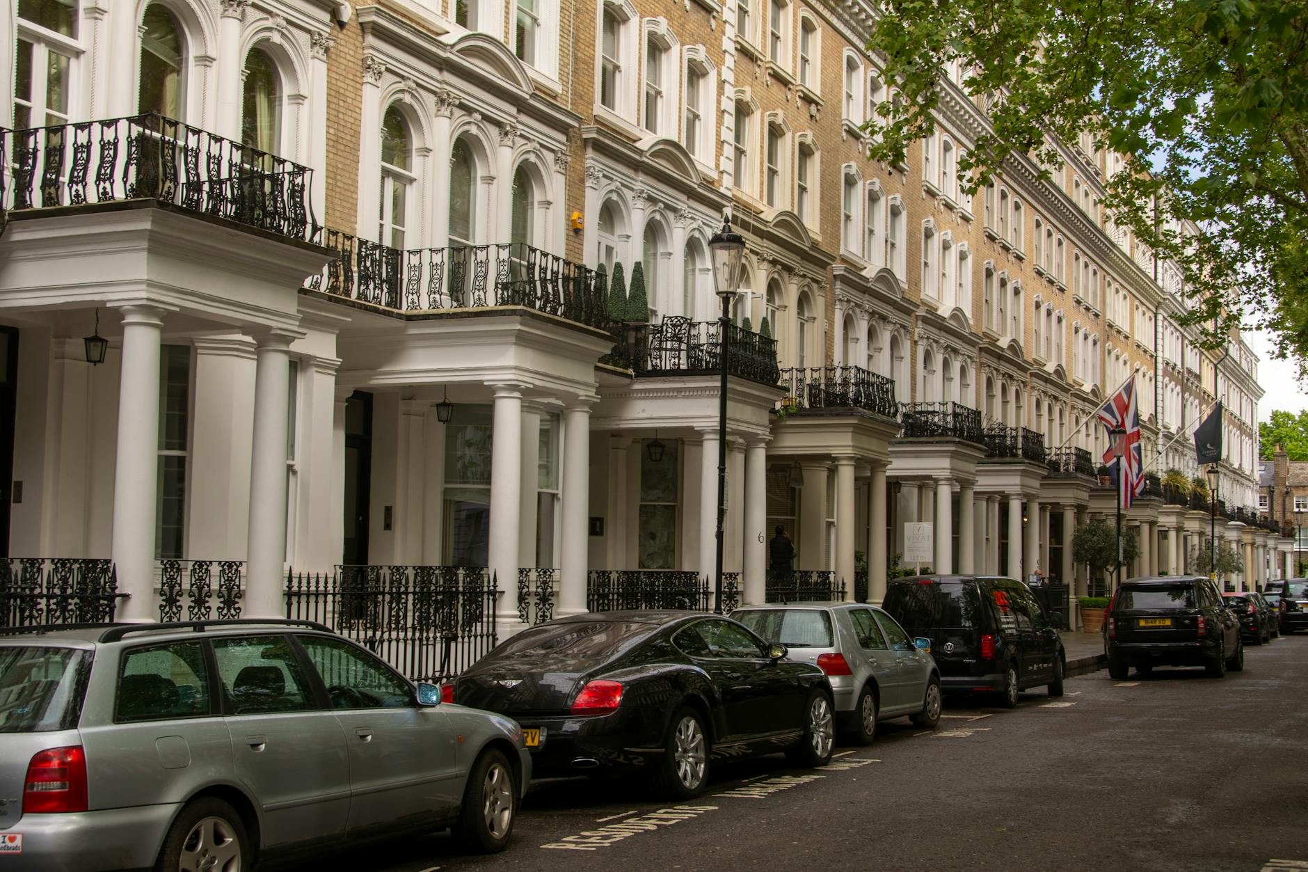 Row of white stucco Georgian townhouses with wrought-iron balconies and a Union Jack flag on a tree-lined London street