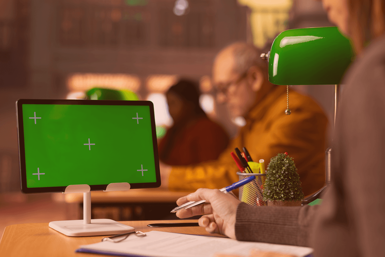 A close-up shot of an office worker's hand holding a pen over a clipboard, with a green screen tablet on a stand in the foreground. In the blurred background, colleagues work under green desk lamps in a warm, professional office environment.