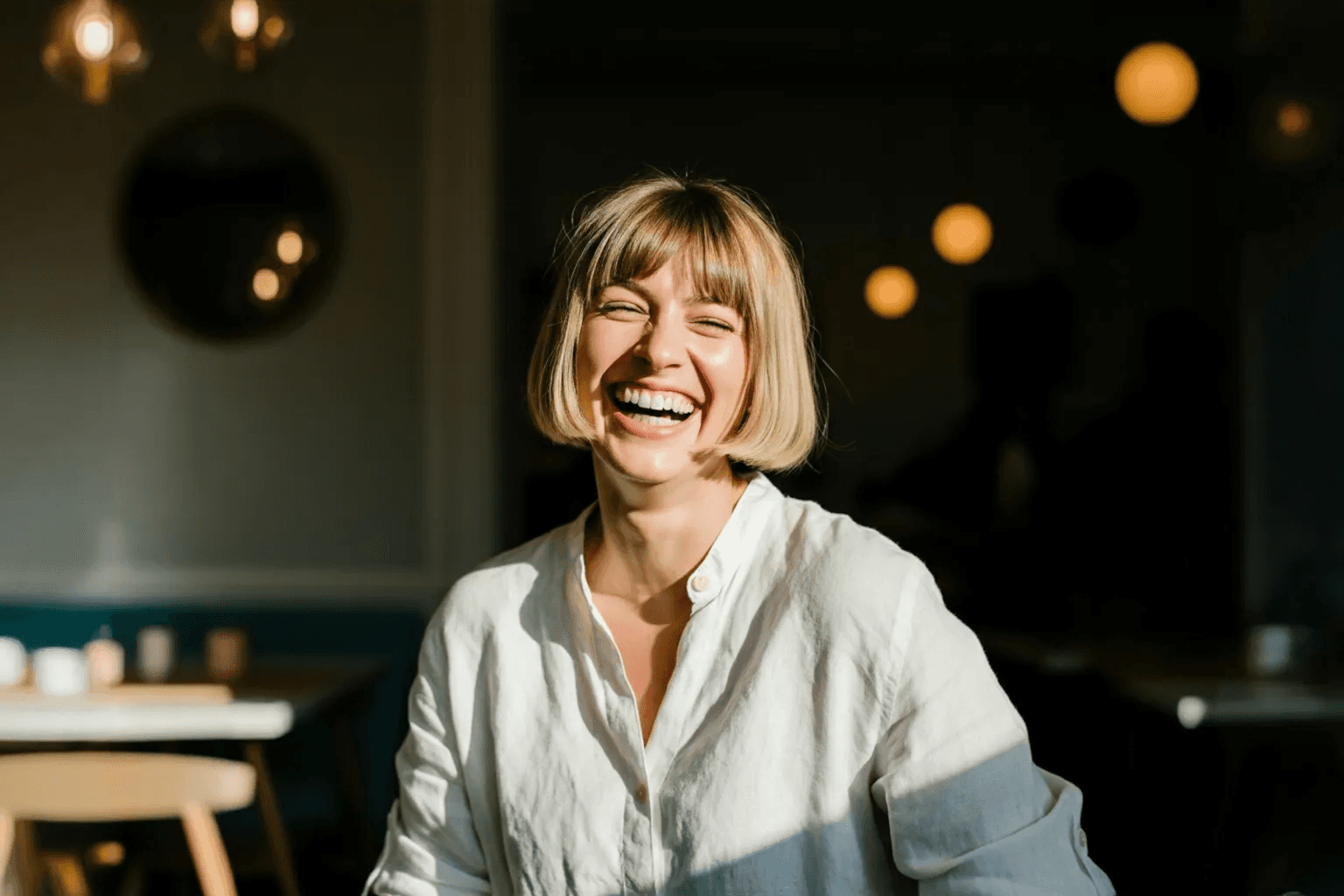 Woman with short blonde hair smiling brightly while sitting in a warmly lit café.