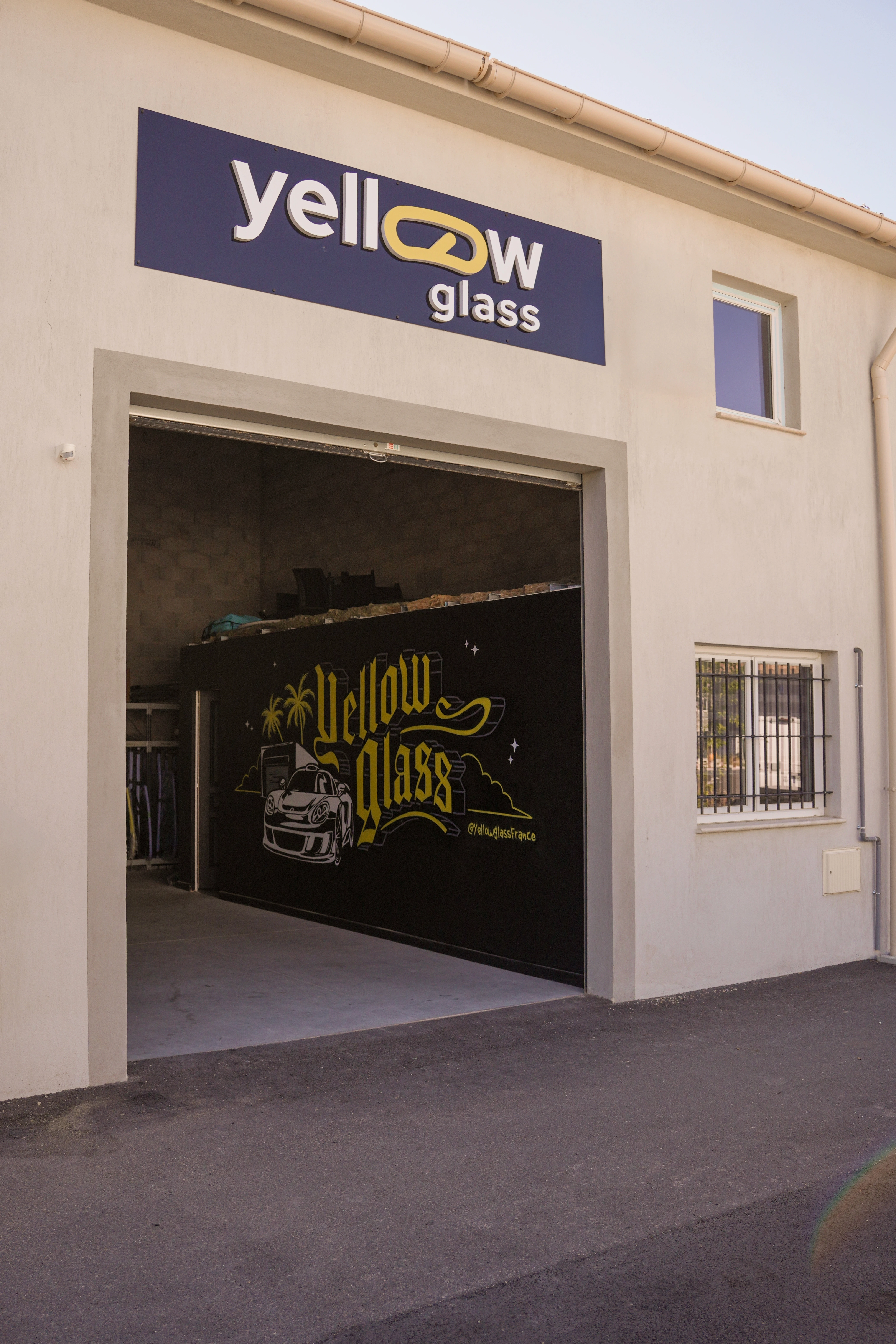 Façade de l'entreprise Yellow Glass avec son enseigne bleue et le logo jaune. La porte de garage est ouverte.