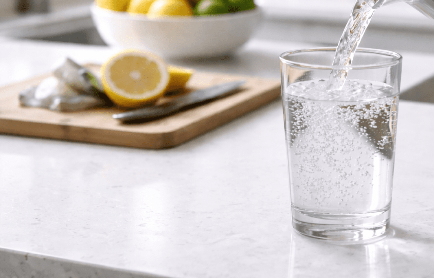 Fresh water being poured into a clear glass with a lemon garnish on a kitchen counter, highlighting product purity.