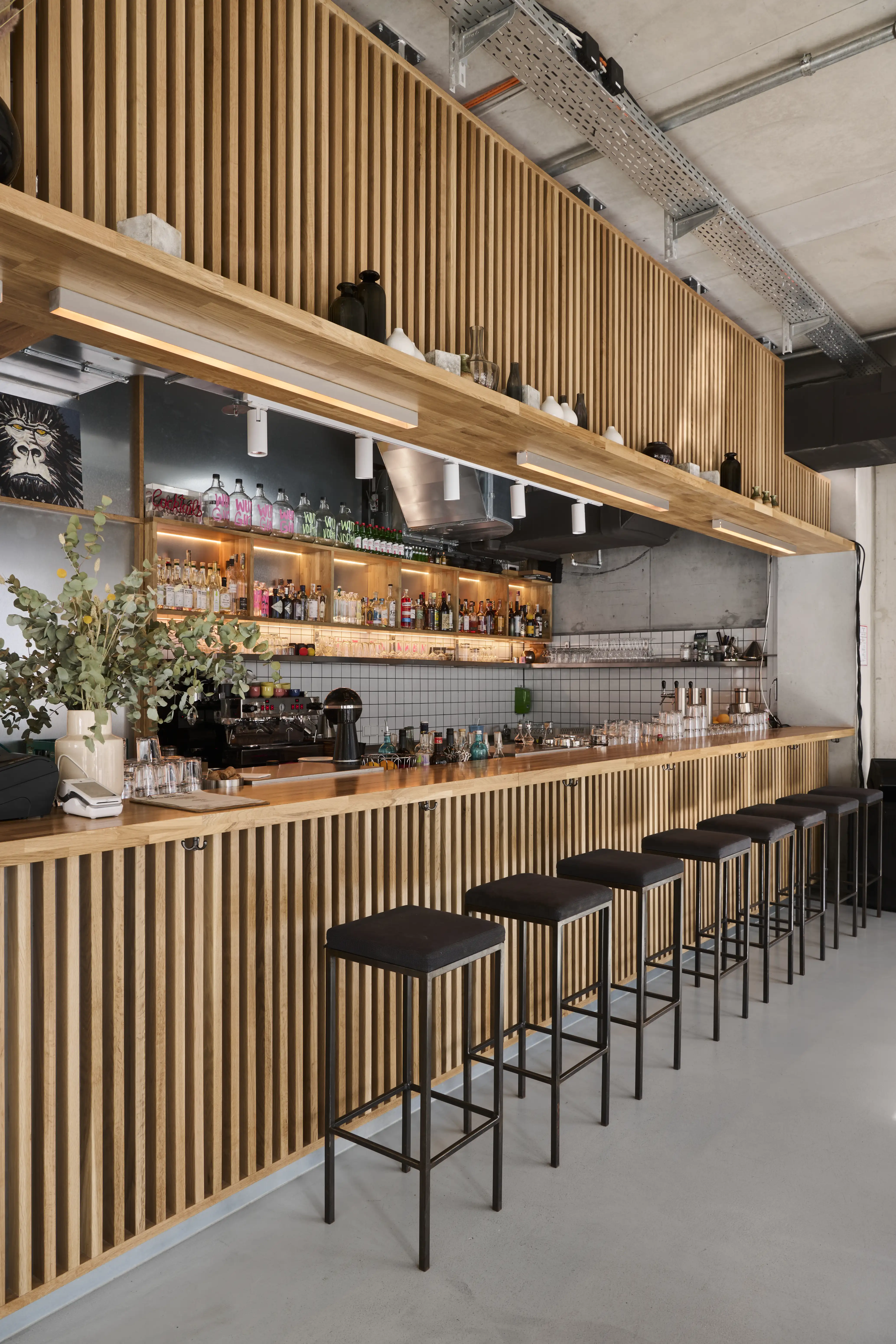 Long bar at PARKER BOWLES & FRIENDS with a vertical wooden slat front, black bar stools, backlit liquor shelves and a wooden canopy with white spotlights.