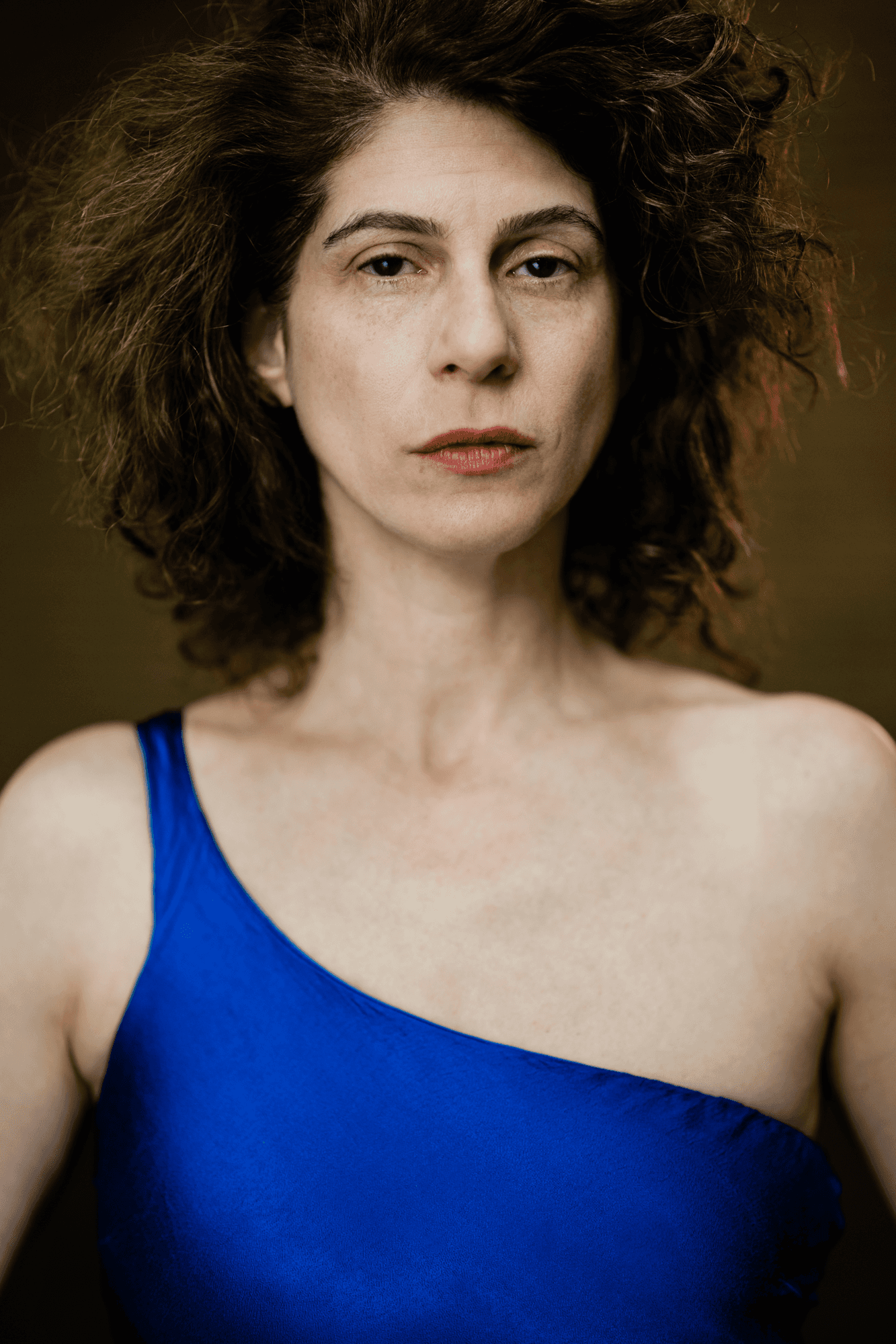 headshot casting photo of Leila Gray, actor, curly-haired woman in blue top, dark studio background.