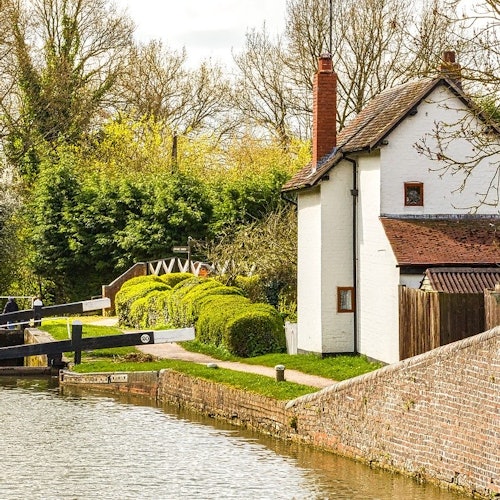 Valkoinen talo, jossa on punainen savupiippu, sijaitsee kanavan sulkuportin vieressä, ympärillään vihreitä pensaita ja puita, aurinkoisena päivänä.