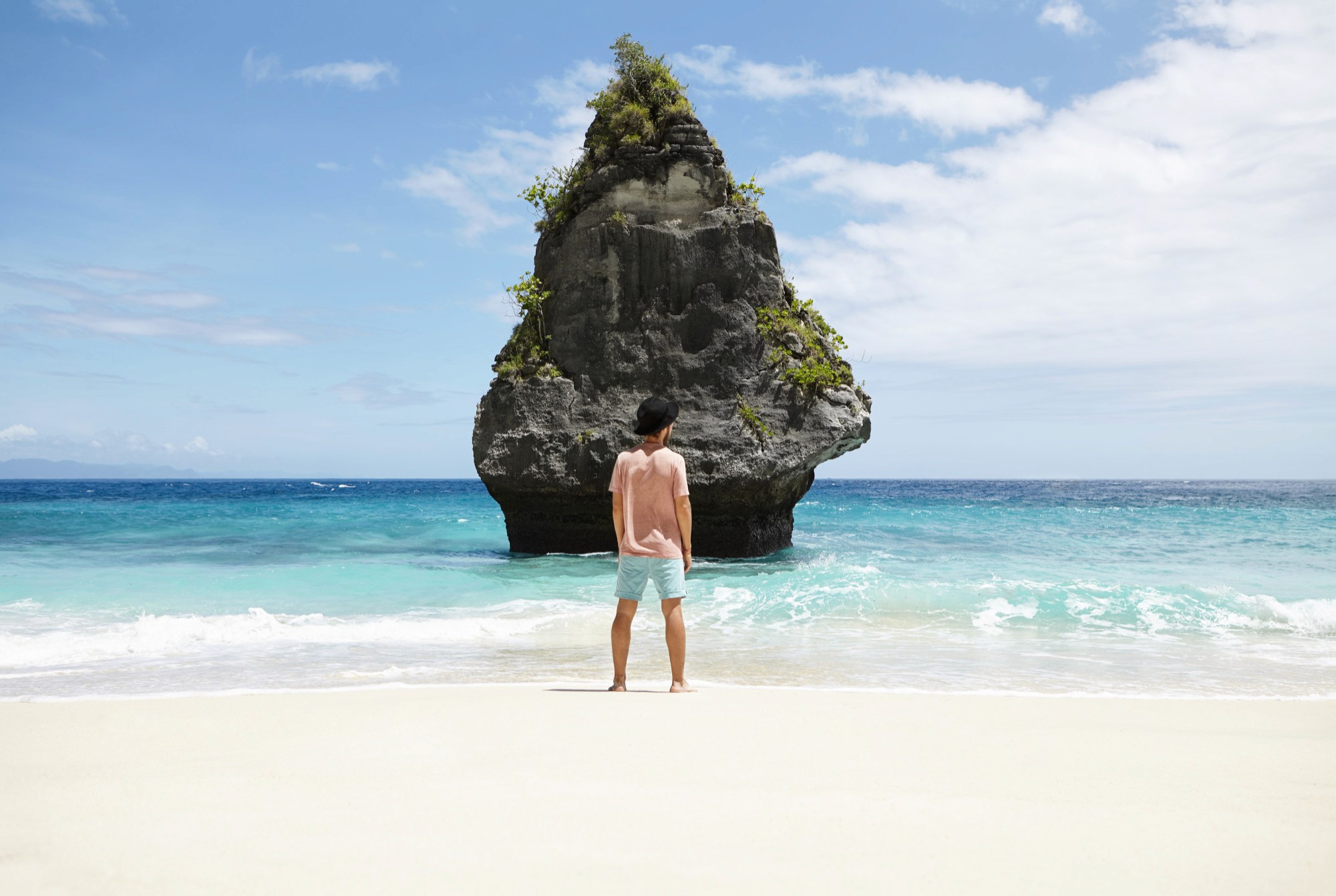 Man standing on a sandy beach facing a turquoise sea and a tall rock island.