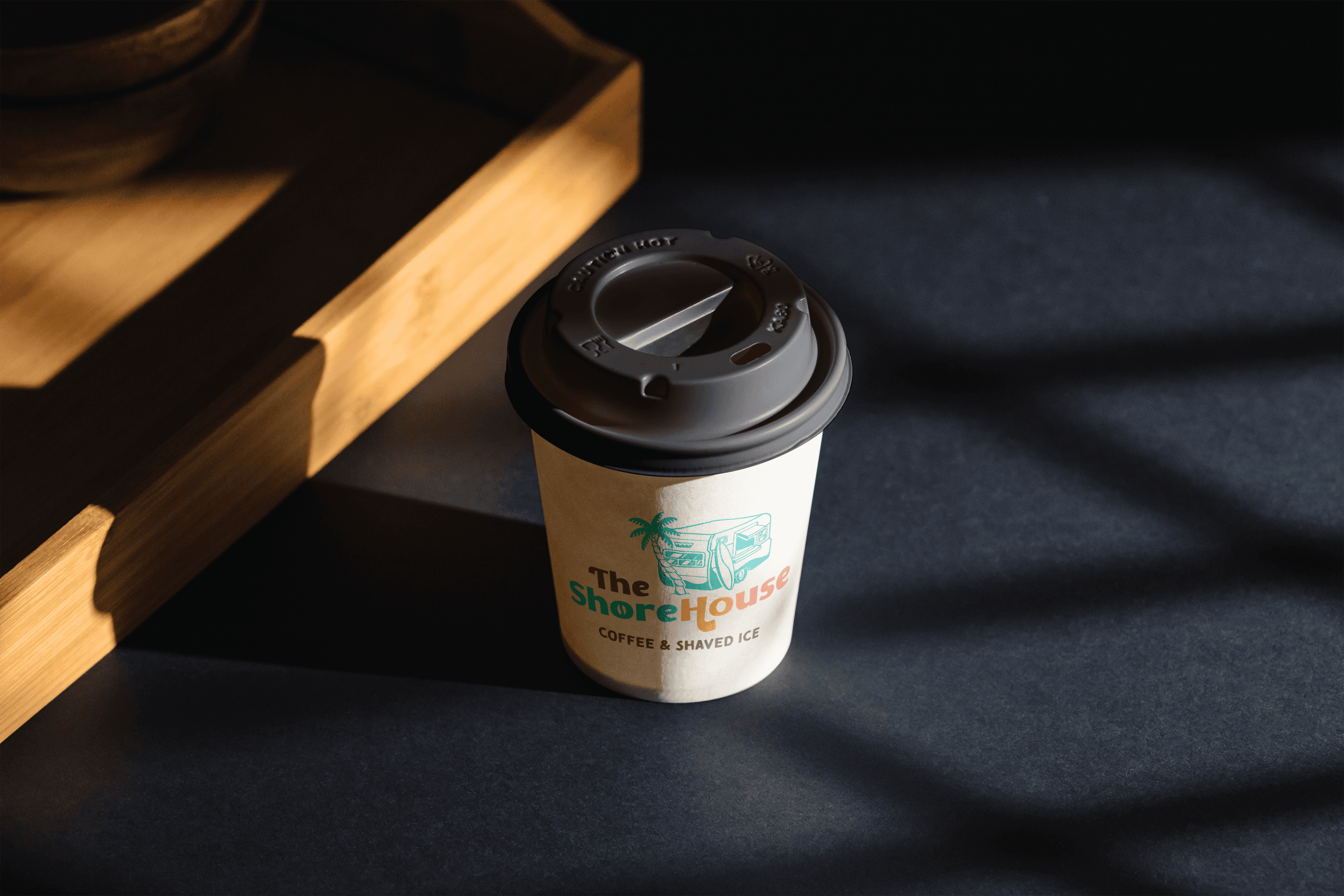 A coffee cup sitting on a table with sun shining on it through a window. The cup has The ShoreHouse Coffee and Shaved Ice logo on it