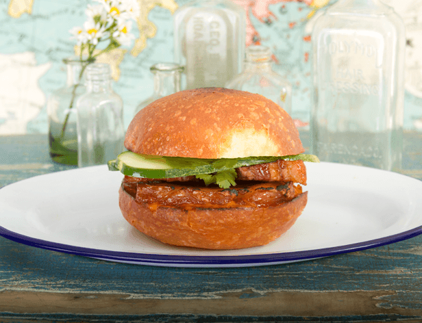 A grilled burger sits on a white plate with a dark blue rim. The burger has a toasted bun, a grilled patty, a slice of cucumber, and green herbs. The plate rests on a blue-green wooden surface. In the background, several glass bottles and a vase with white flowers are visible. The background is out of focus and does not show any map or detailed objects.