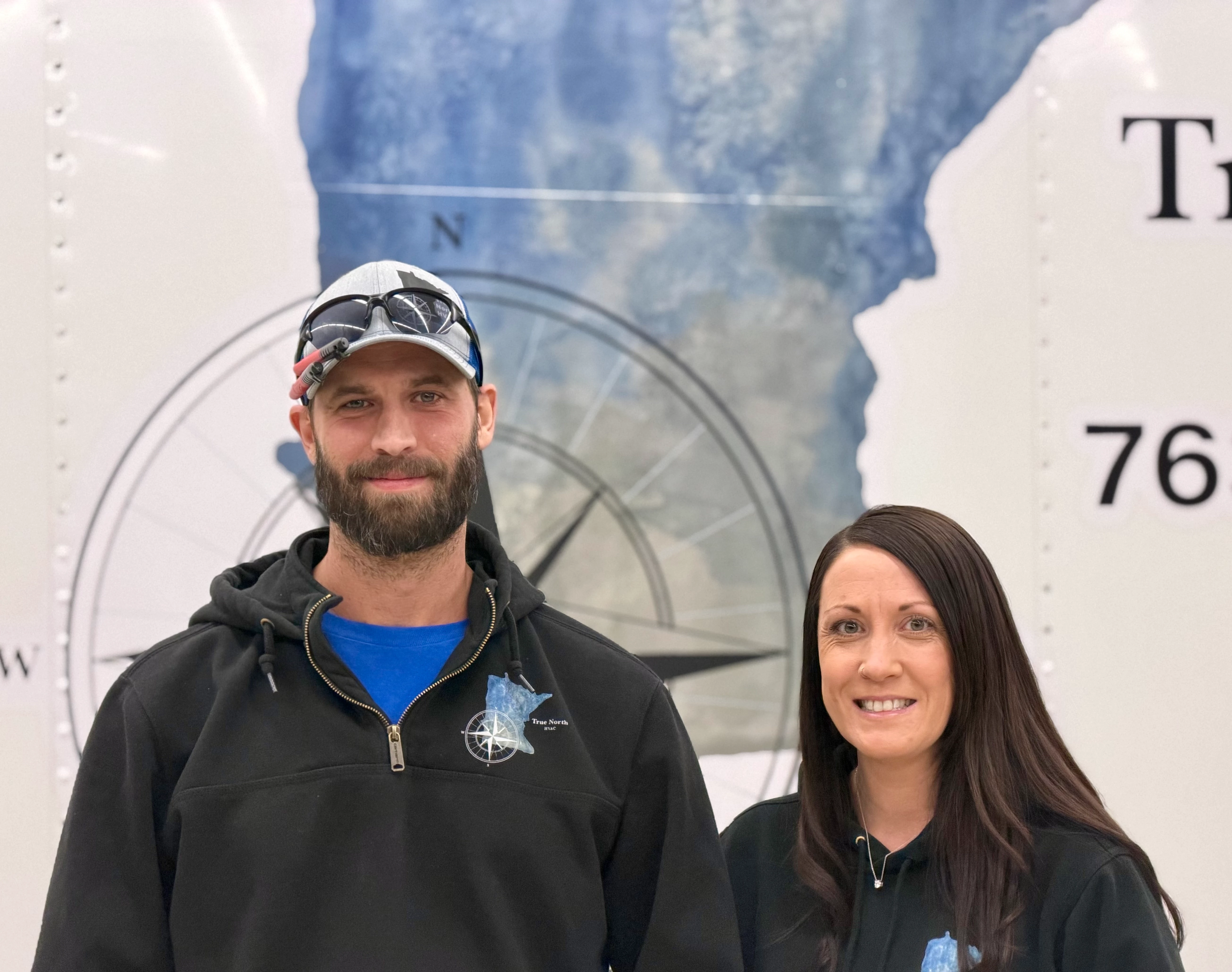 A young person with curly hair smiles at the camera while sitting in front of a laptop against a dark background.