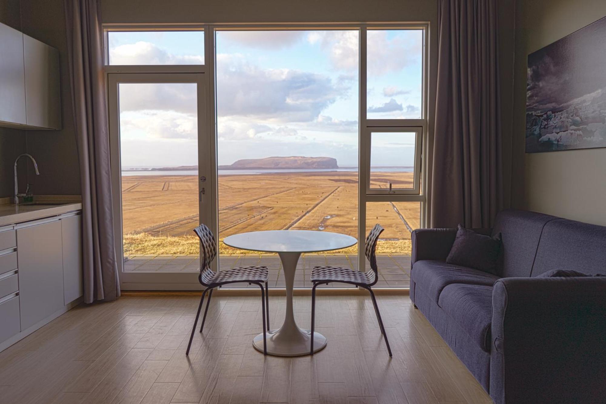 Inside of a studio apartment with a kitchenette, table and two chairs and a small sofa with a view outside a large window of Icelands mountains and views