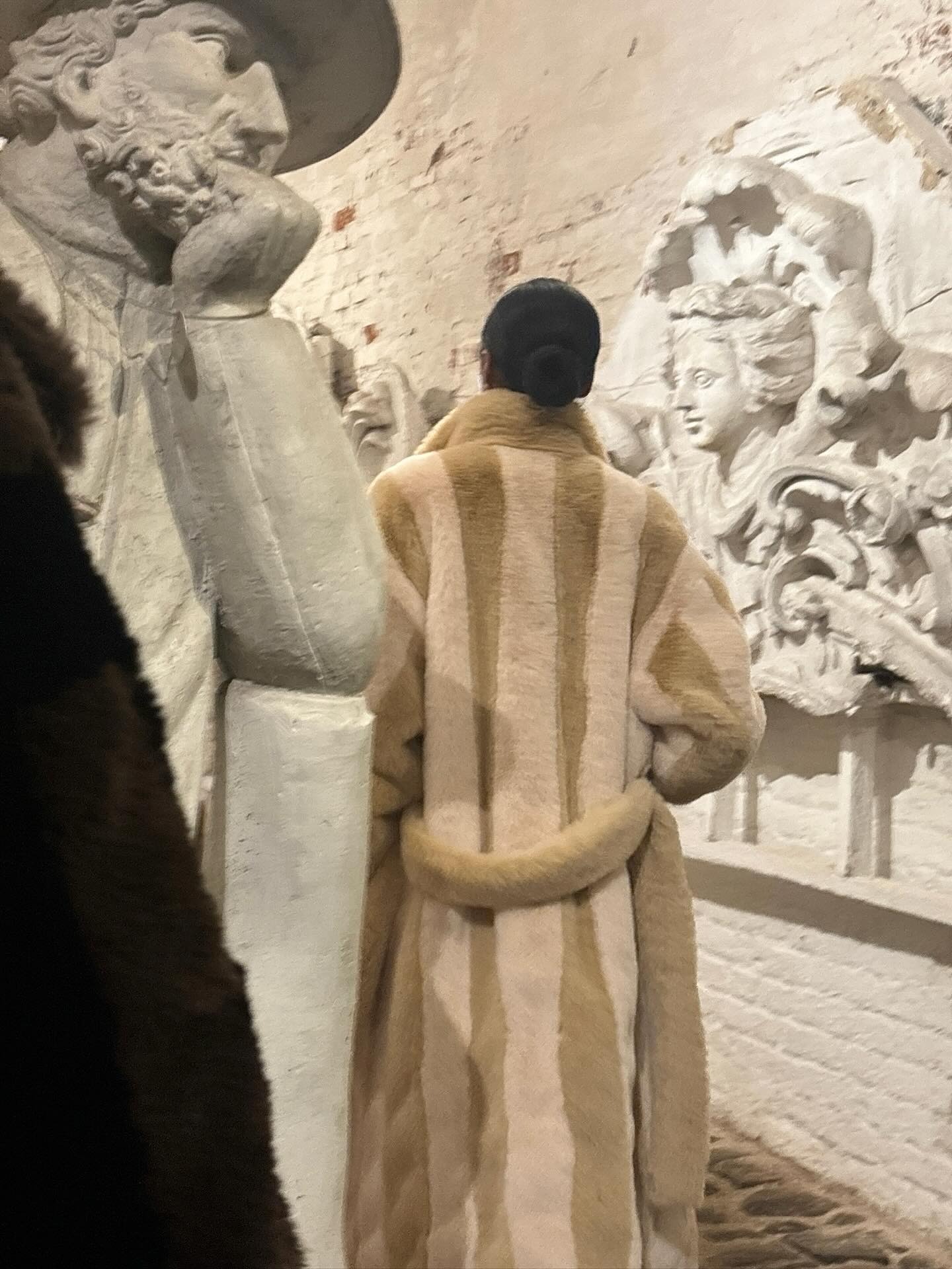 Entre 2 statuts, une femme porte un manteau en fausse forrure beige/marron