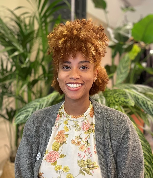 A person standing indoors in front of large leafy green plants, creating a natural, softly lit backdrop. They have short, tightly curled light‑brown hair and are wearing a light floral top with pastel flowers, layered under a grey cardigan. A small pin is attached to the cardigan, and the overall setting feels warm and relaxed, similar to a studio or office space with indoor plants.