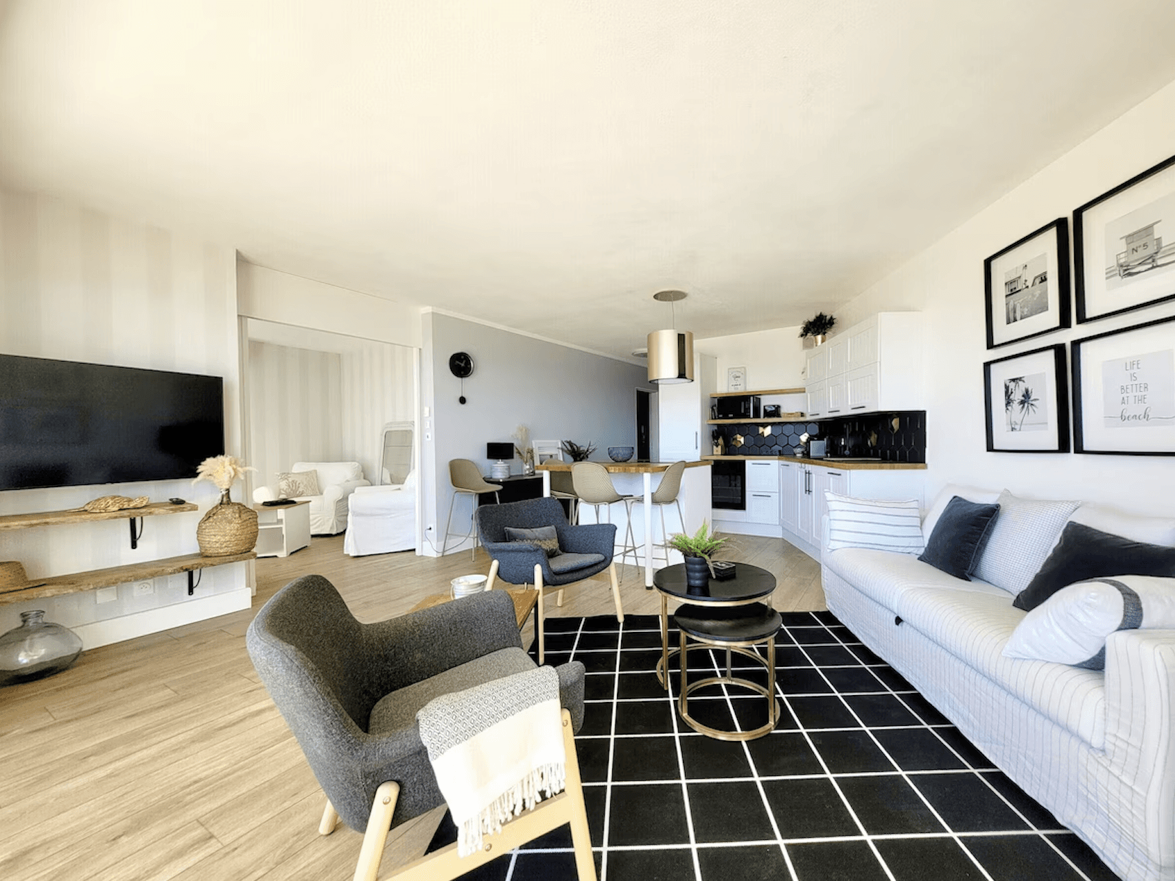 Spacious modern apartment with a kitchen island, black chairs, and stainless steel appliances. A cozy living area with armchairs and plants in the background.