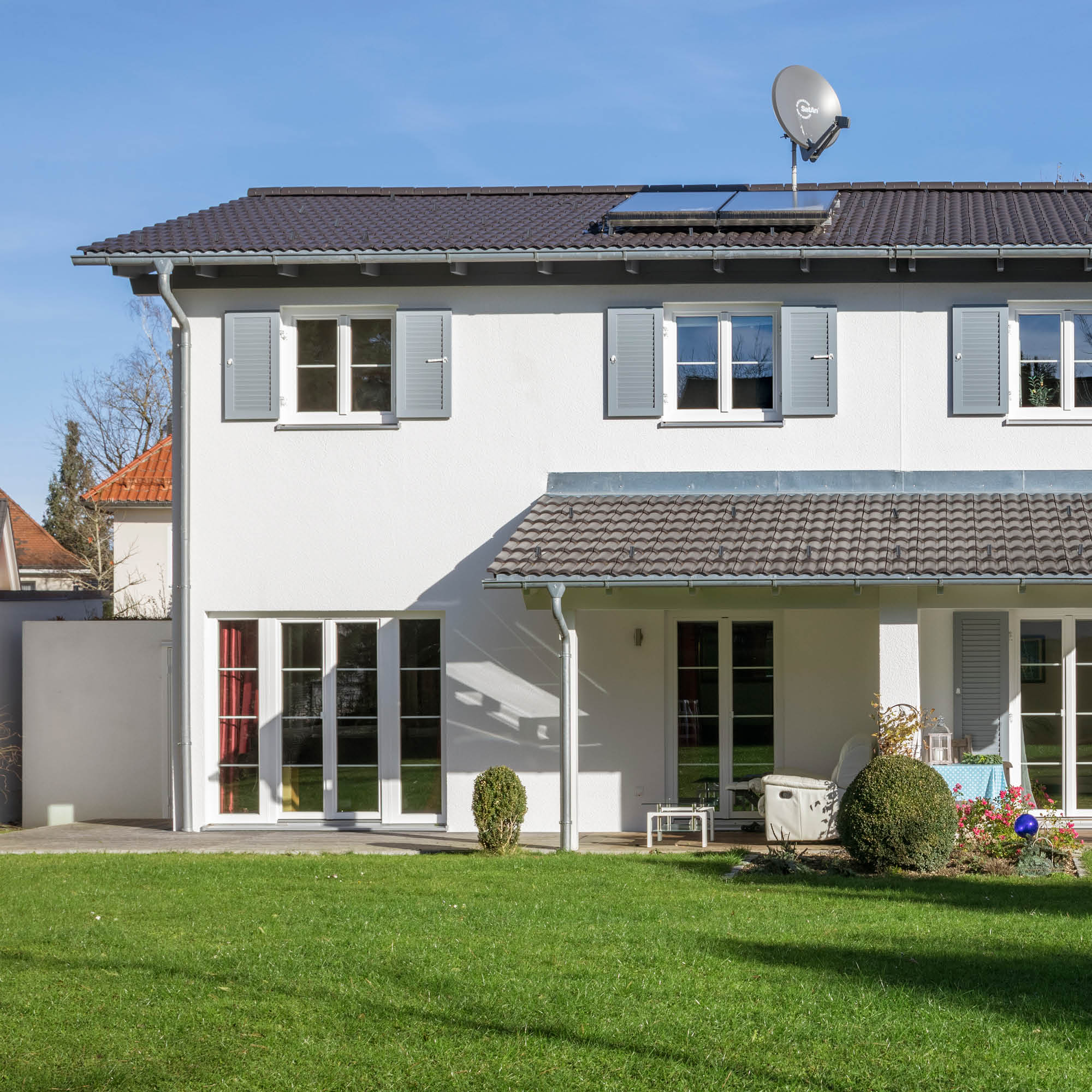 Fassade eines modernen Hauses mit weißem Putz, grauen Dachziegeln, Satellitenschüssel, Solarmodulen und einem gepflegten Garten.