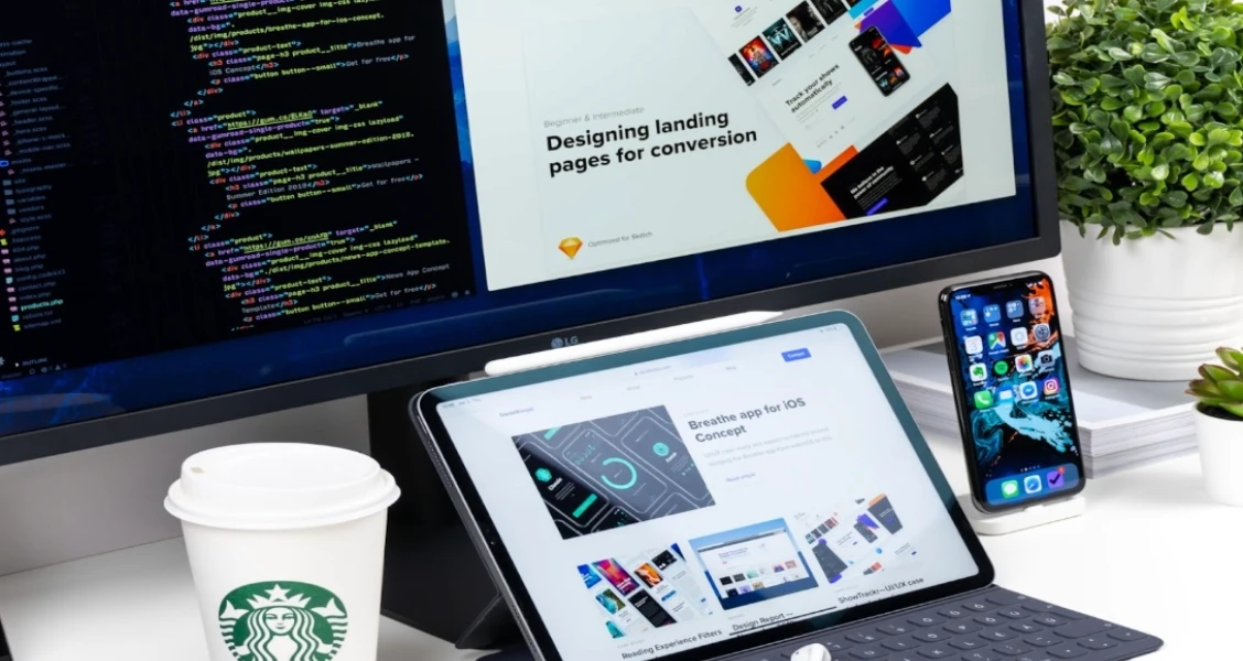 Workspace setup featuring a desktop monitor displaying code and a landing page design, a tablet showing a website interface, and a smartphone on a stand, arranged on a white desk with a coffee cup and a small plant—illustrating a multi-device landing page design environment.