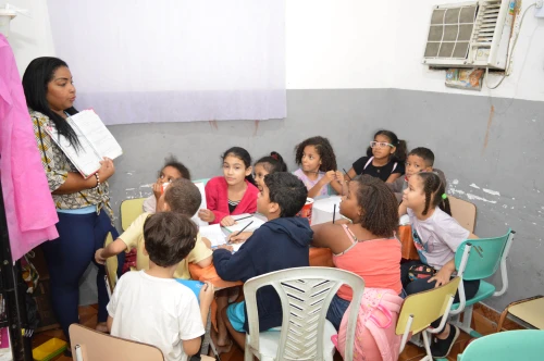 Crianças em círculo prestando atenção na professora, em uma sala de aula simples com ar-condicionado.