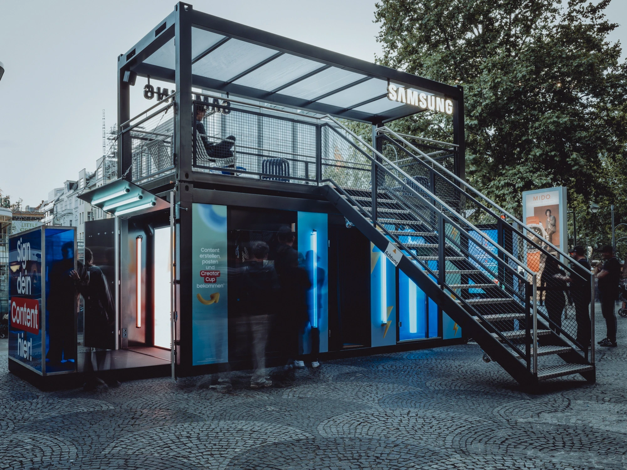 Two-storey black container event module for SAMSUNG x TIKTOK with blue-lit walk-in booths, external steel staircase and roof terrace with Samsung lettering.