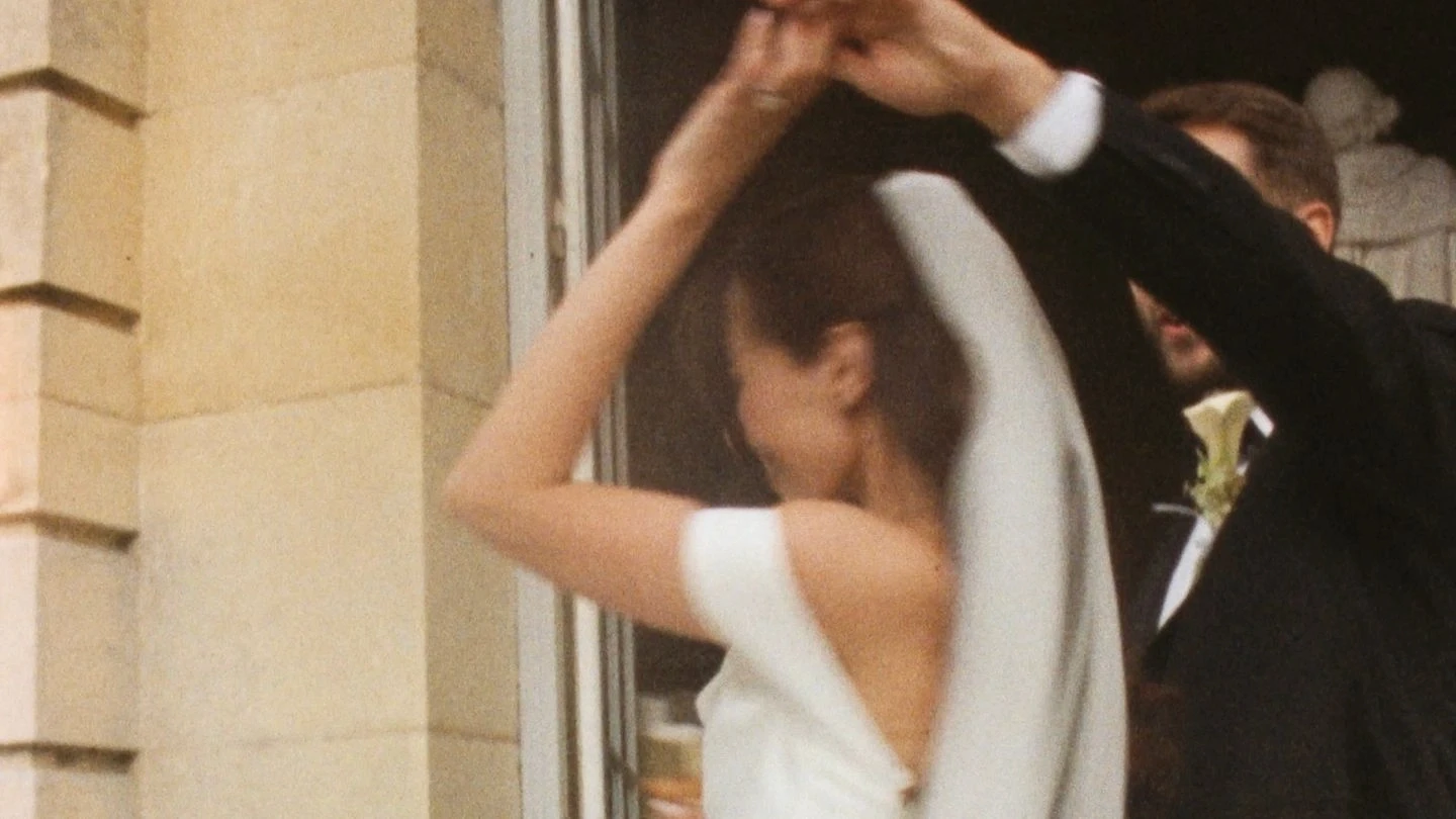A bride in an off-the-shoulder white gown and a groom in a suit share a joyful dance at an elegant wedding venue entrance.