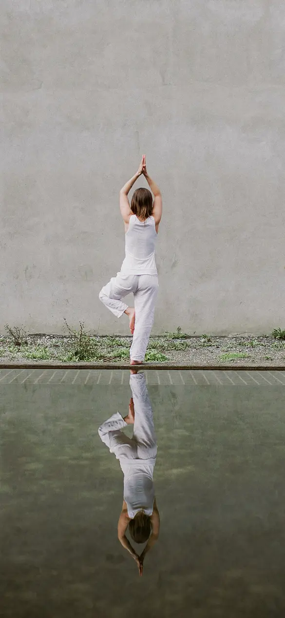A person practices yoga in a serene outdoor setting, standing near a tranquil reflecting pool with a minimalist stone wall in the background, embodying wellness and recovery at a premium spa-like environment.