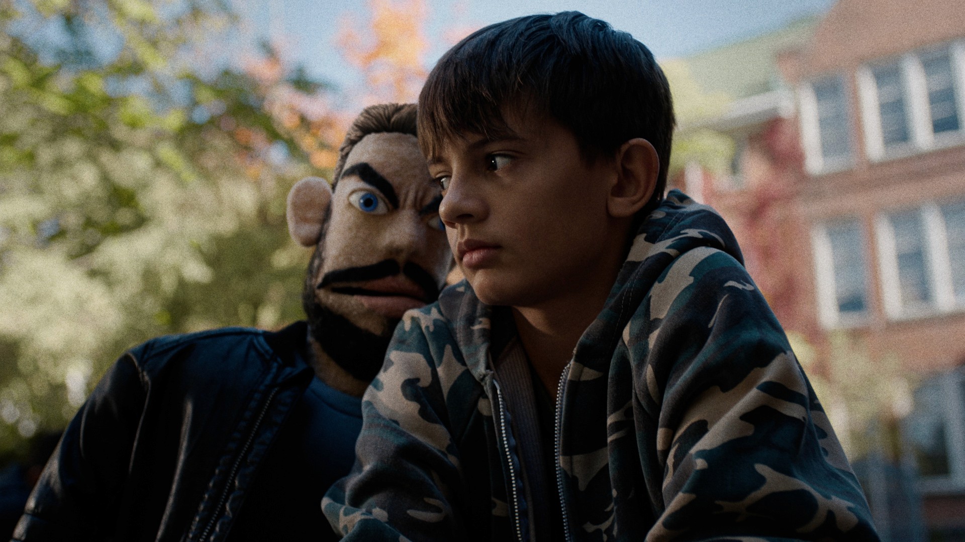A young boy in a camo hoodie looks pensive while sitting outdoors. Beside him, a puppet with exaggerated eyebrows and leather jacket intimidates him.
