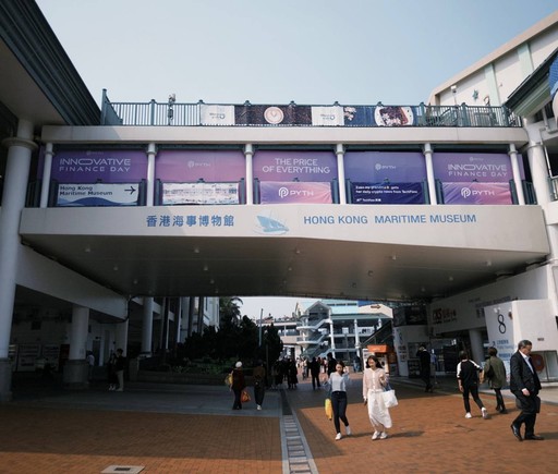 Entrance walkway to the Hong Kong Maritime Museum, featuring large purple banners promoting “Innovative Finance Day” and “The Price of Everything” by Pyth Network. People walk underneath the overpass in a sunlit plaza with signs and storefronts visible on both sides.