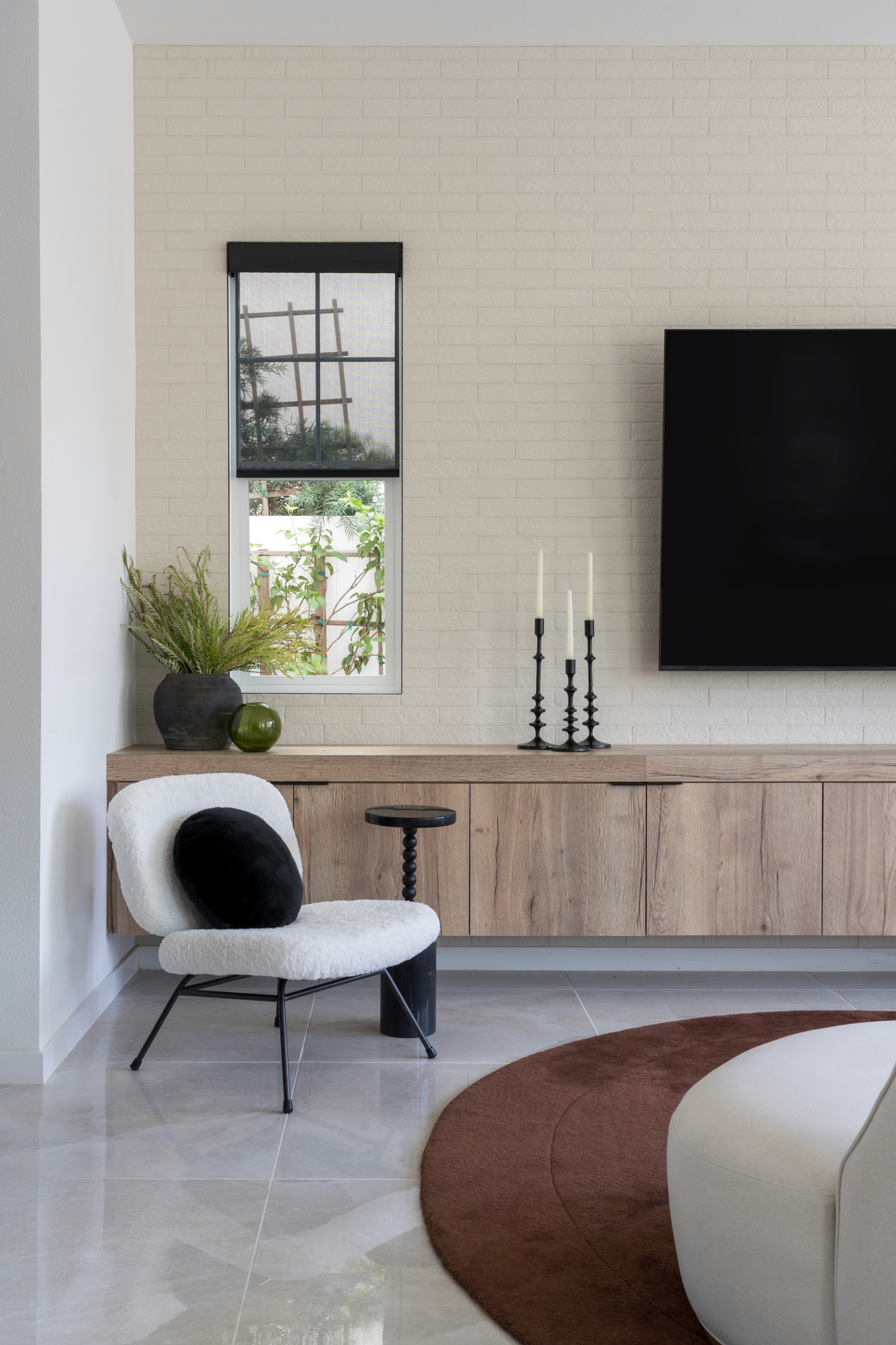 Living room with warm brown oval rug, white accent chair, neutral wood built-in media console, and white brick accent wall by Jubilee Interiors in Arcadia, California