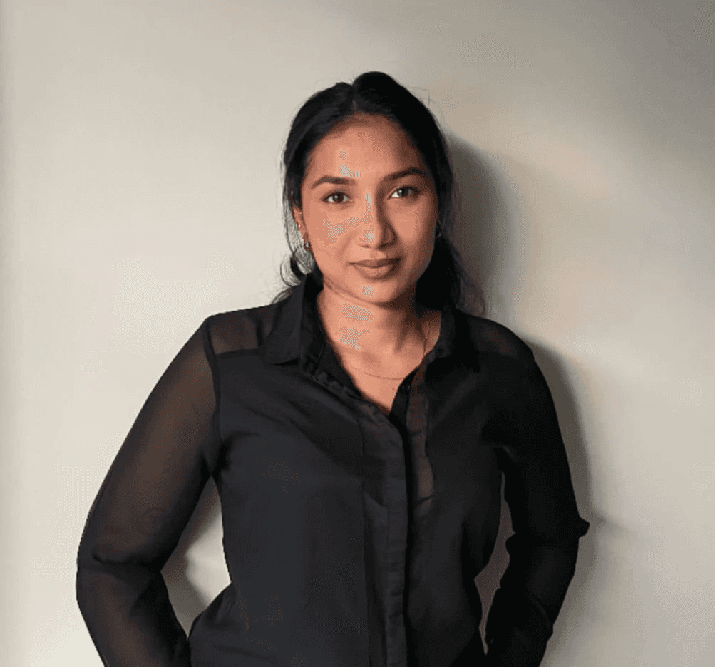 Young adult wearing a navy coat and white turtleneck, looking directly with a neutral expression. The background is plain and light-colored.