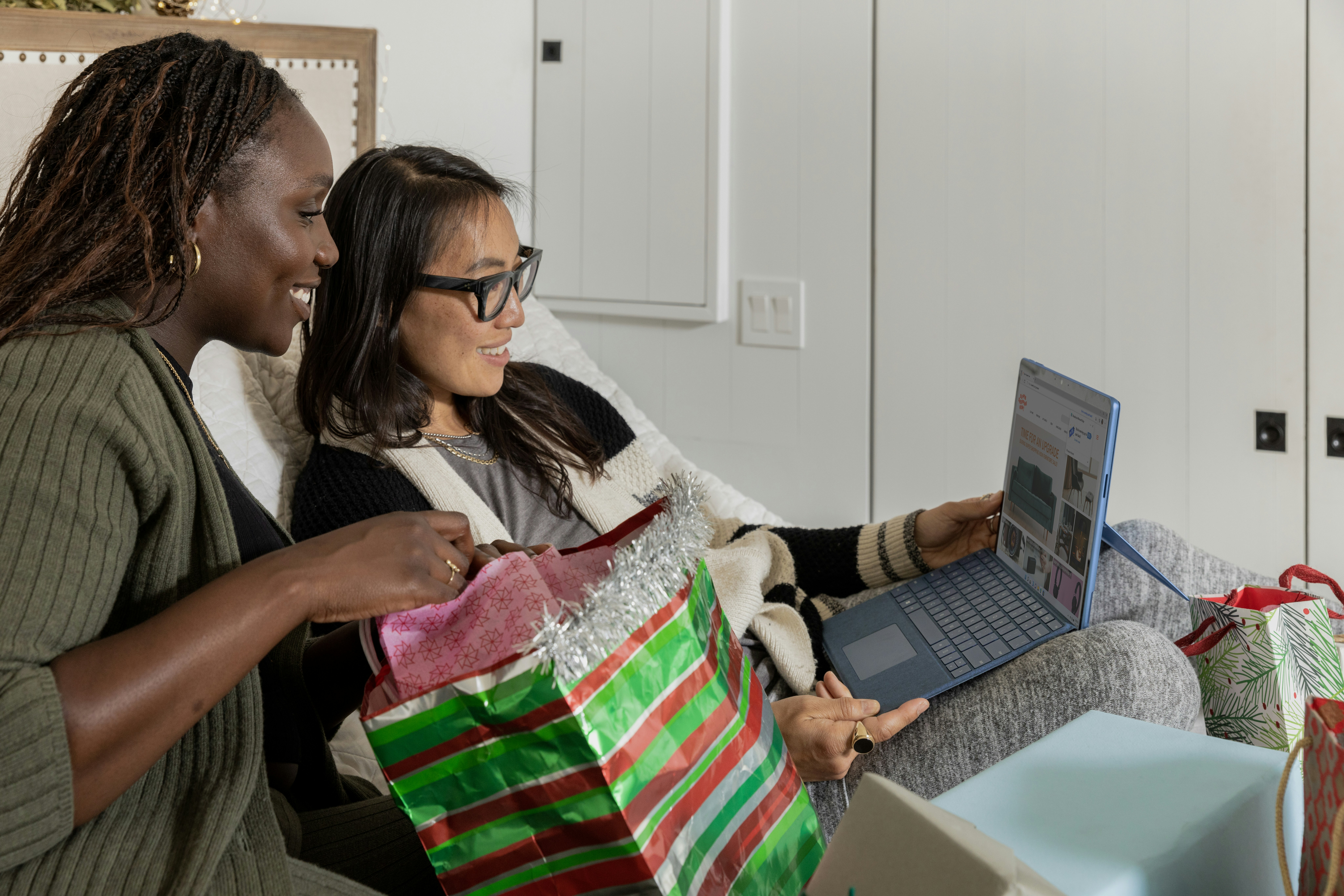 Two women sitting on a couch surrounded by presents, using a laptop and enjoying each other's company.