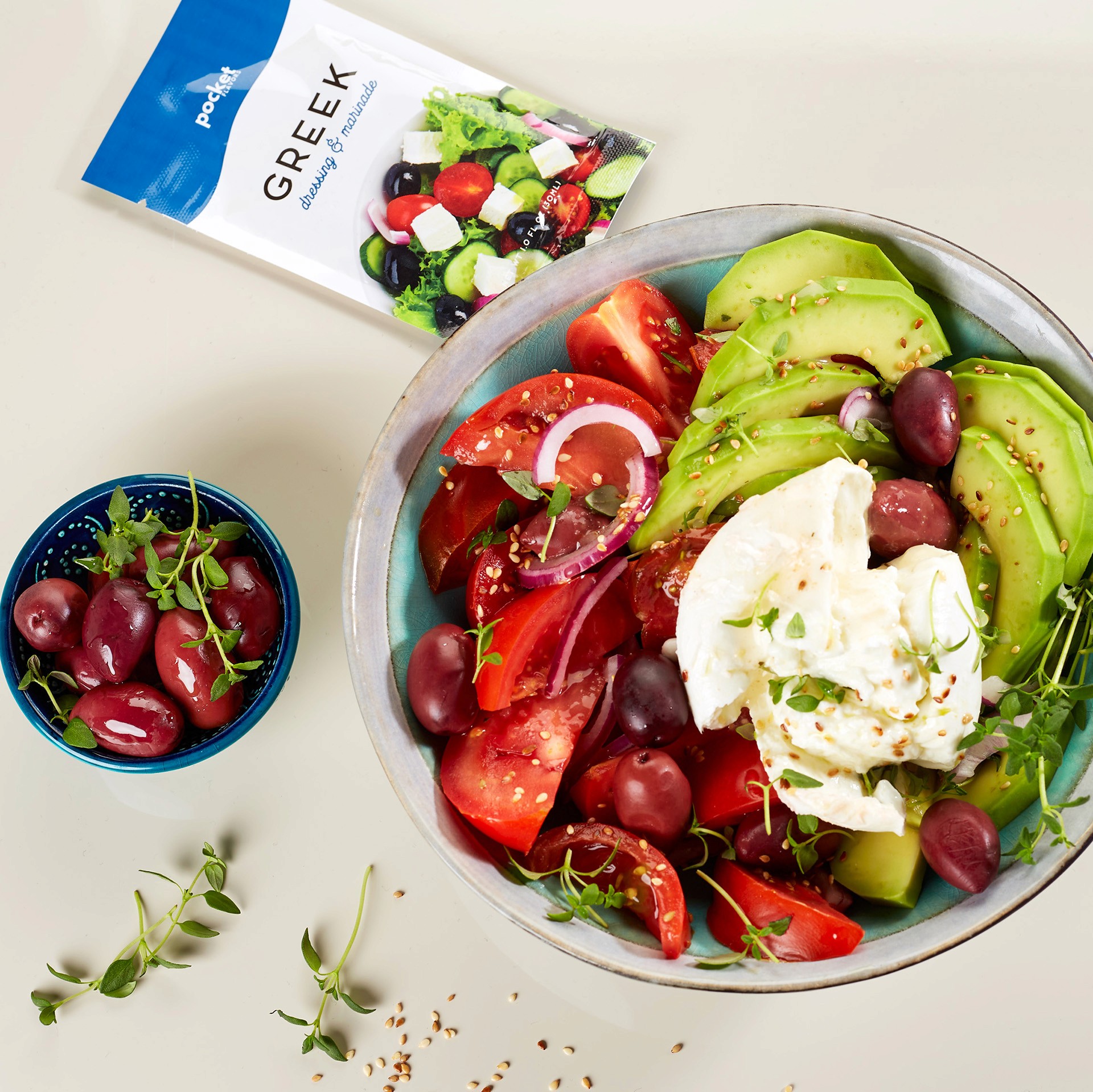 A colorful Greek salad featuring sliced avocado, fresh tomatoes, red onion, black olives, and a dollop of creamy cheese, with a package of salad seasoning and a small bowl of olives nearby.