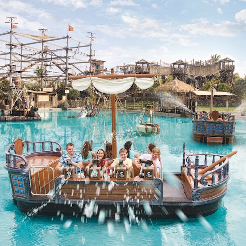 Kinderen en volwassenen op een piratenthema-boottocht met waterkanonnen, ingericht in een pretpark met een touwenparcours op de achtergrond.