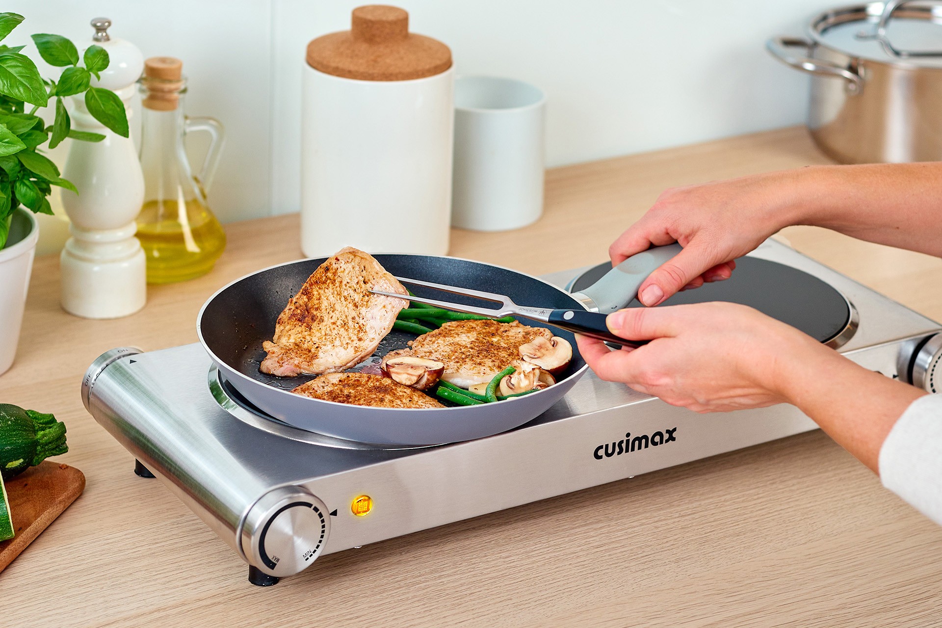 A person is using kitchen tongs to flip seasoned chicken breasts in a skillet on a sleek, stainless steel Cusimax portable electric burner, surrounded by various kitchen ingredients like herbs, olive oil, and zucchini, indicating a cooking session in a modern kitchen setting.