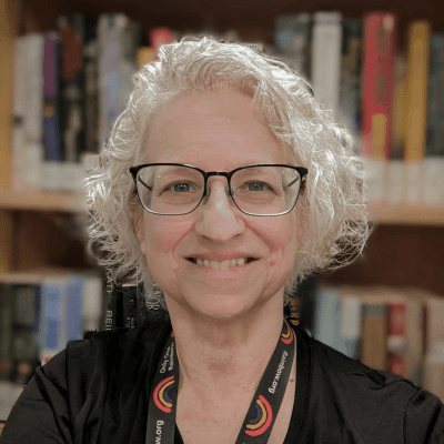 Portrait of author Nancy Seeger smiling at the camera, wearing glasses and a black top, with bookshelves softly blurred in the background.