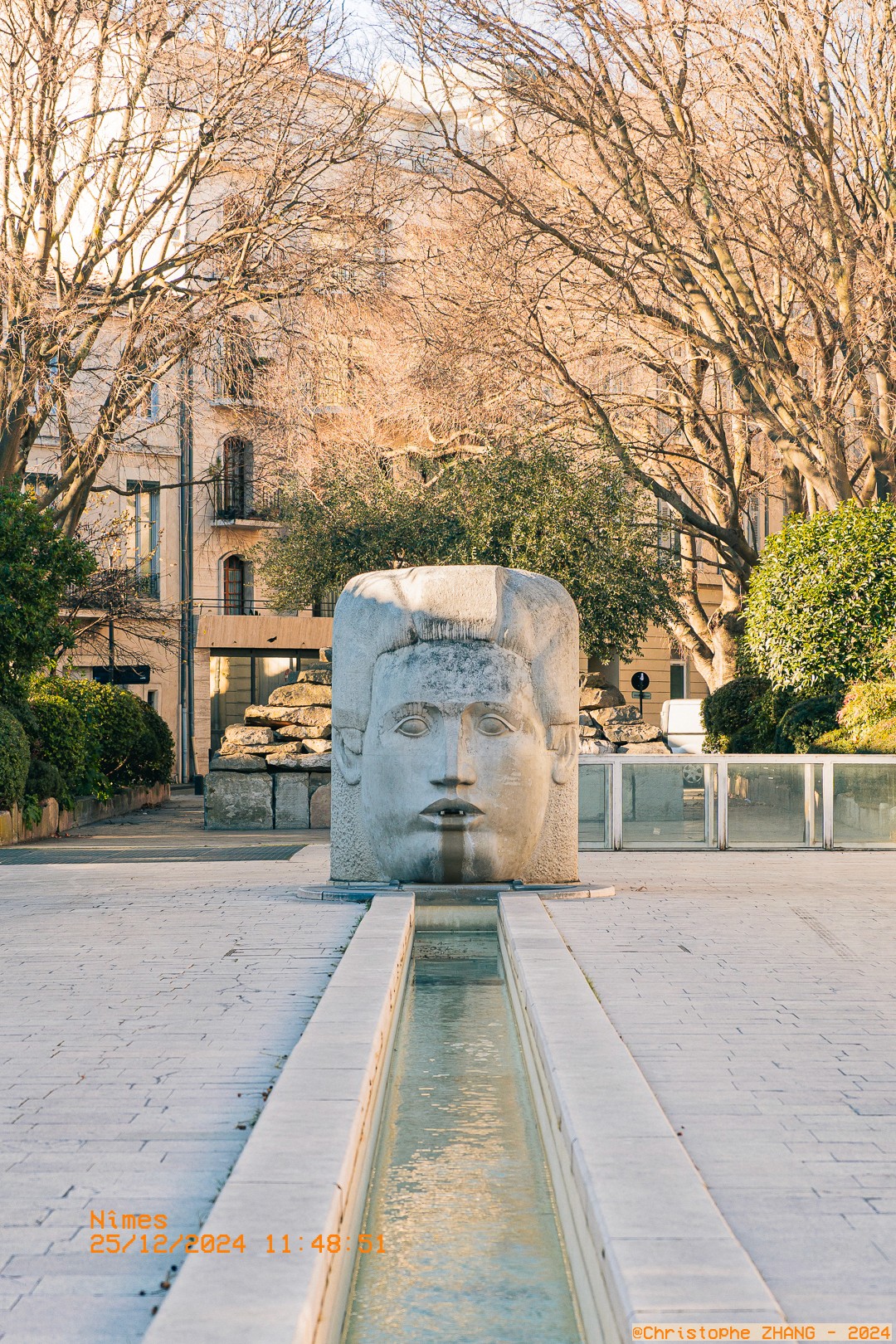 Perspective frontale de la fontaine Némausus à Nîmes, entourée d’arbres et d’éléments architecturaux.