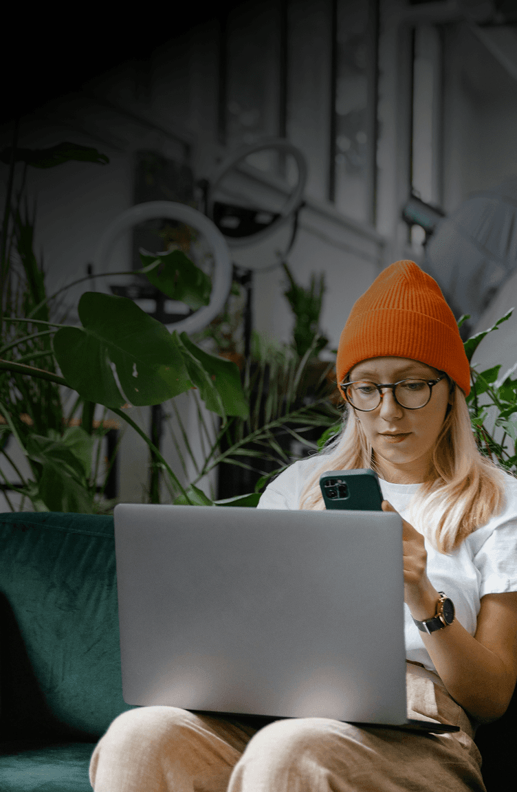 A young woman with long blonde hair, wearing an orange beanie and glasses, sits on a green couch focused on her smartphone while a laptop is open in front of her. The background features indoor plants and a modern workspace, creating a cozy and productive atmosphere.