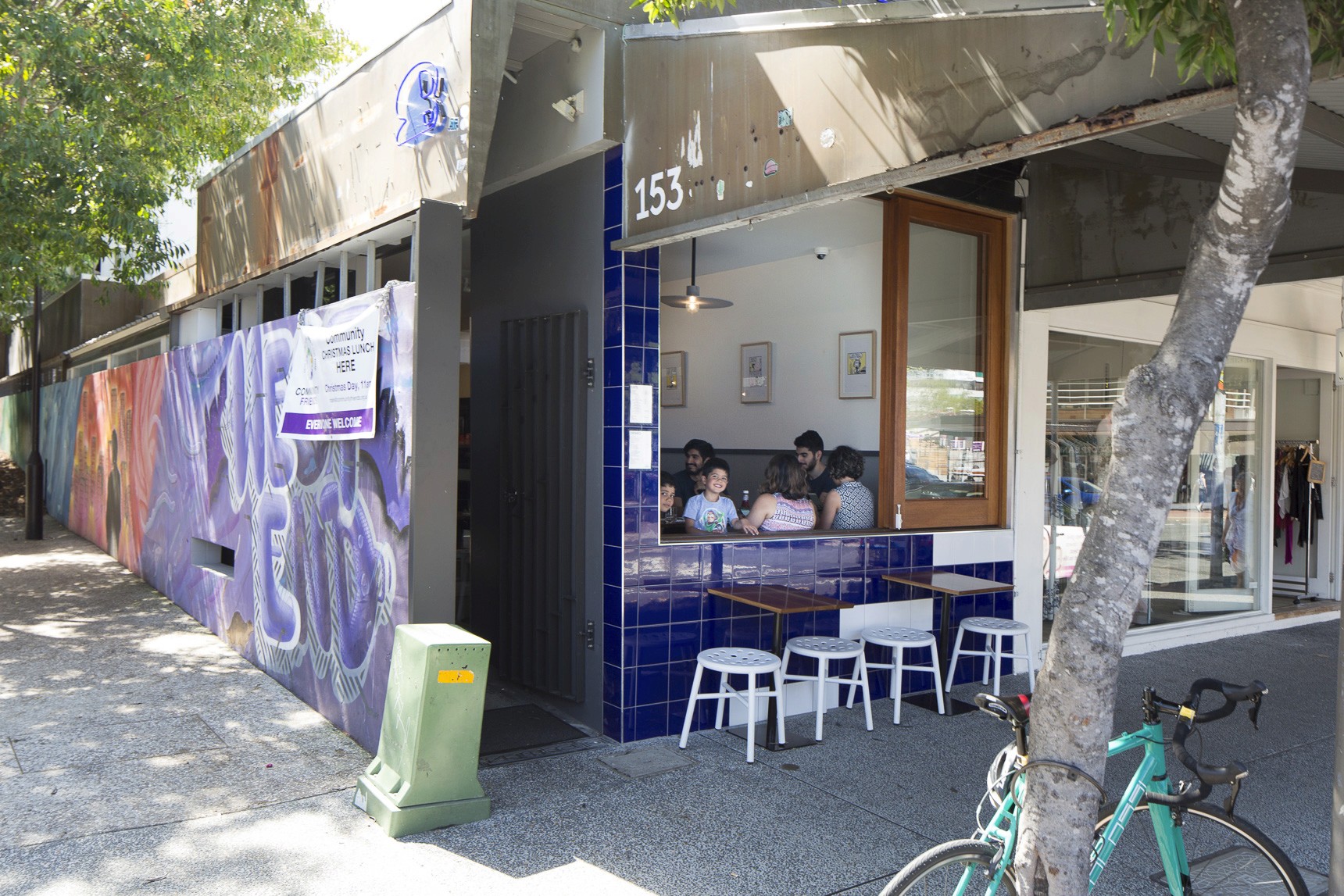Street-facing exterior of Ben’s Burgers West End featuring blue tiled window seating, white stools along the footpath, mural-lined side wall, and diners visible through an open service window under dappled tree shade.