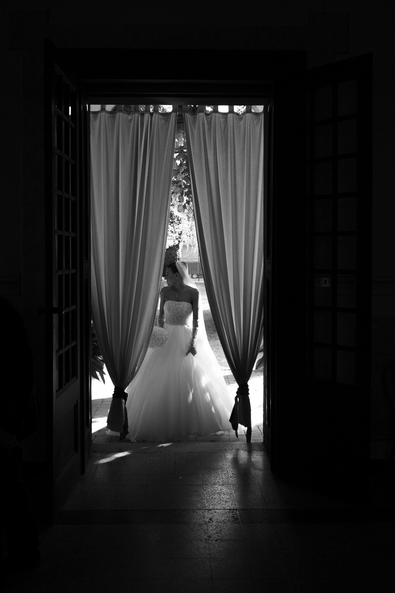 Sposa in abito bianco visto da dietro, tra le tende drappate dell'ingresso di Villa Ferrari, in bianco e nero