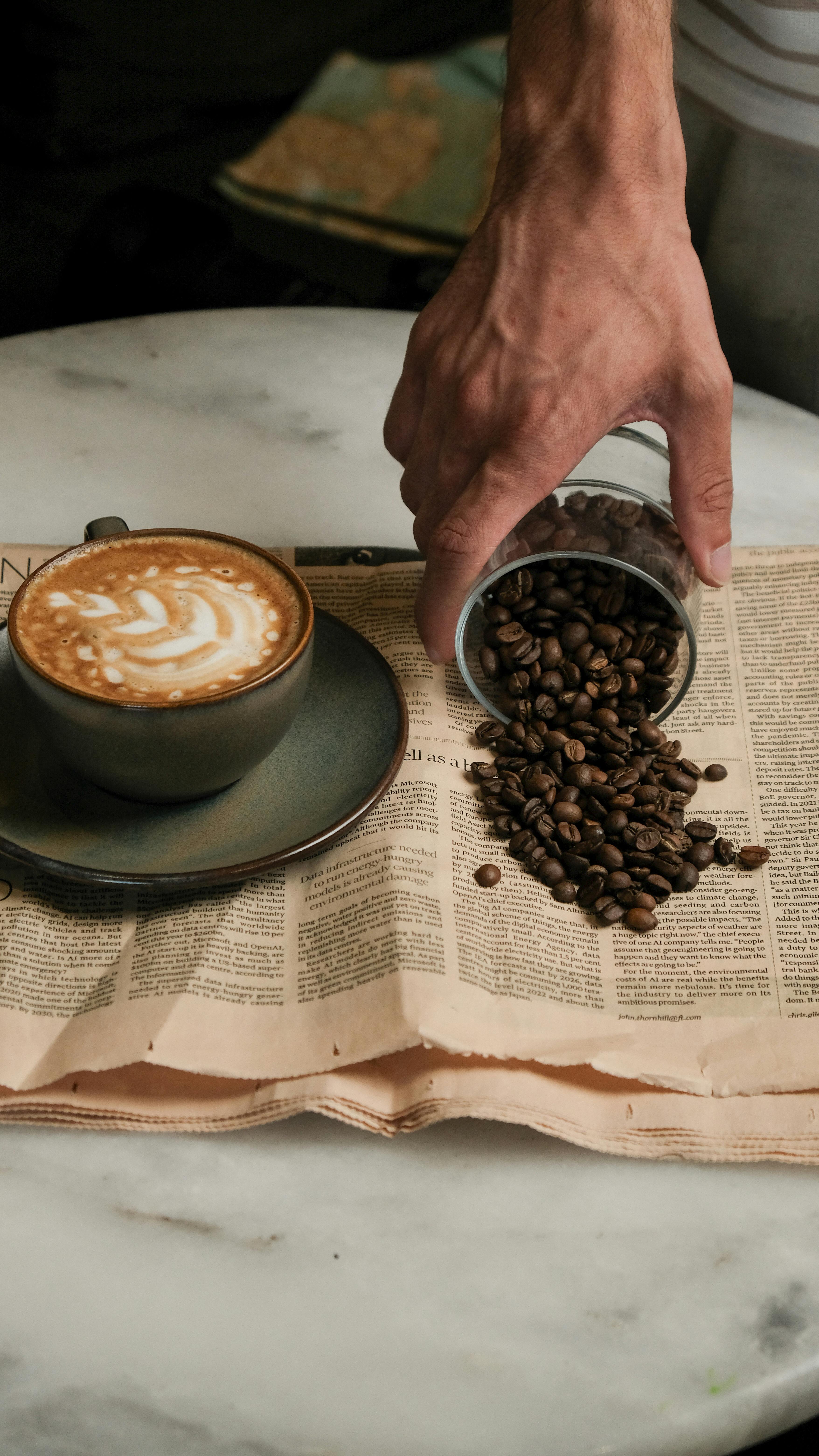 A hand pours roasted coffee beans from a glass jar onto an open newspaper beside a ceramic cup of cappuccino with latte art, all resting on a round marble table.