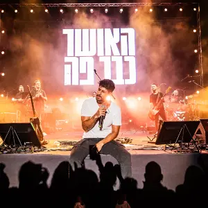 עידן עמדי בהופעה חיה על הבמה, יושב ושר מול הקהל תחת הכיתוב המואר 'האושר כבר פה'. צילום הופעה מרגש עם תאורה חמה ועשן במה, צולם על ידי הצלמת שירי עציץ Shiriz Photography.