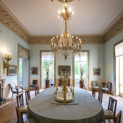 Elegant dining room with a large chandelier, an oval table with a decorative centerpiece, surrounded by chairs and paintings on the walls.