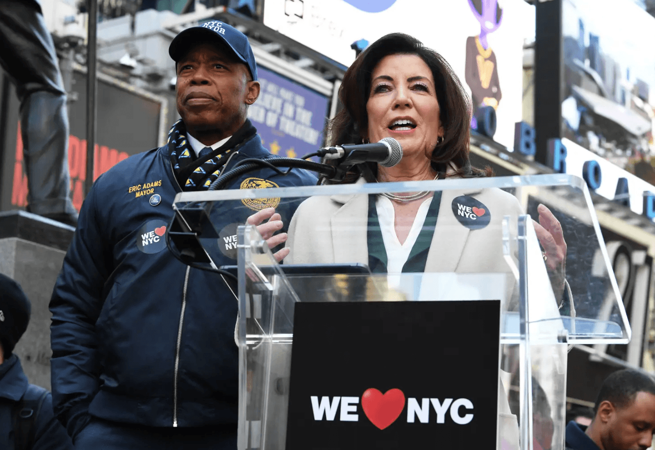 Mayor Eric Adams and Governor Kathy Hochul stand behind a speaker lectern with the We Love NYC logo in Times Square