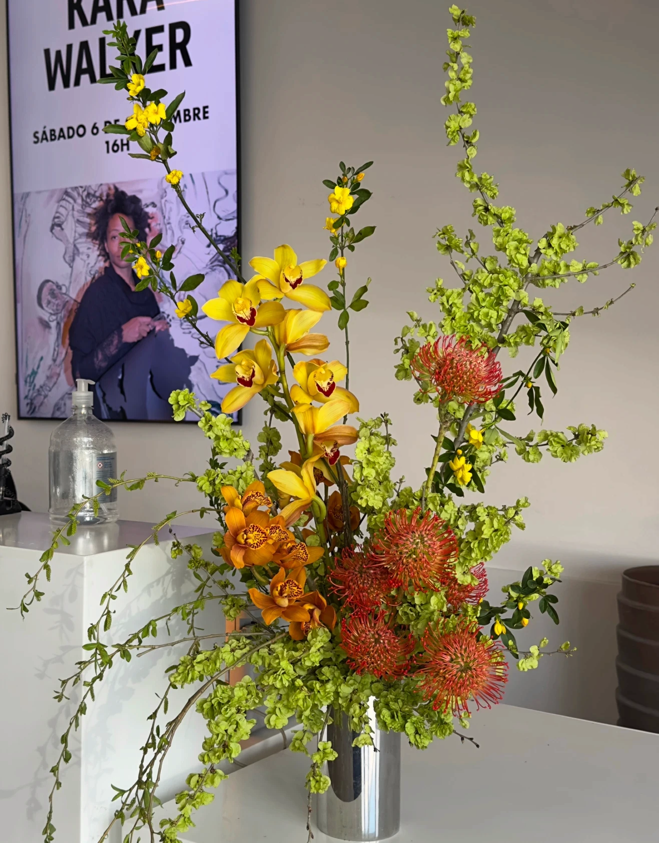 Arreglo de orquídeas y proteas en jarrón plateado sobre mostrador en una galería.